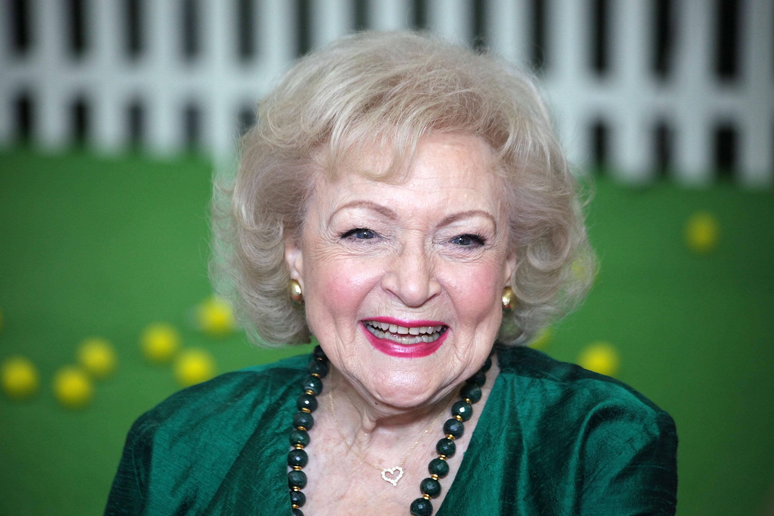 Betty White 桌布 壁紙 — 一位快樂的老年女性，金色捲髮，穿著綠色天鵝絨上衣，在風格化的綠色和白色背景前燦爛微笑。