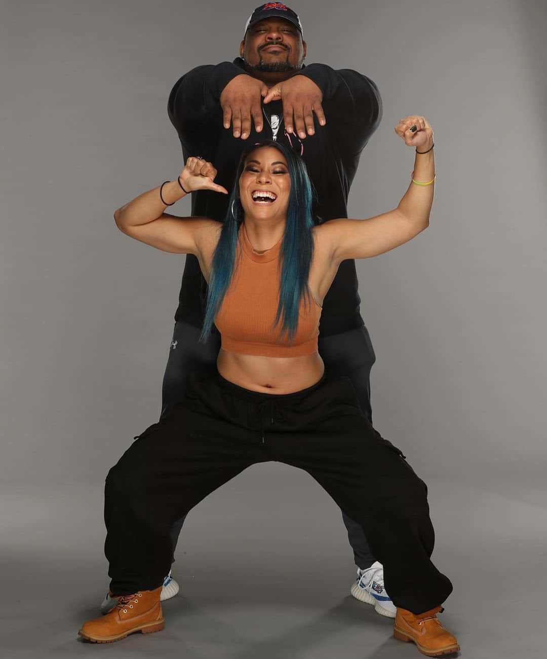 WWE 桌布 壁紙 — 一位快樂的藍髮女子穿著橙色上衣擺出俏皮姿勢，而一名戴帽子的男子站在她身後，雙手放在她的肩膀上。