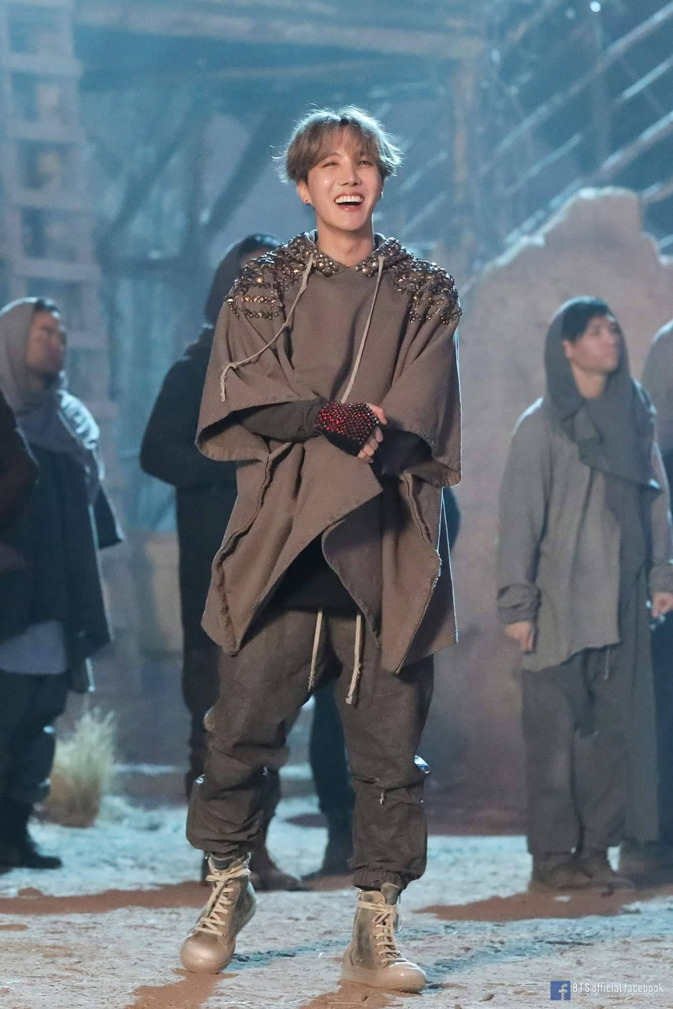 jhope 桌布 壁紙 — 一位快樂的表演者穿著粗獷的棕色服裝，在燈光昏暗的舞台上燦爛地笑著，背景有工作人員。