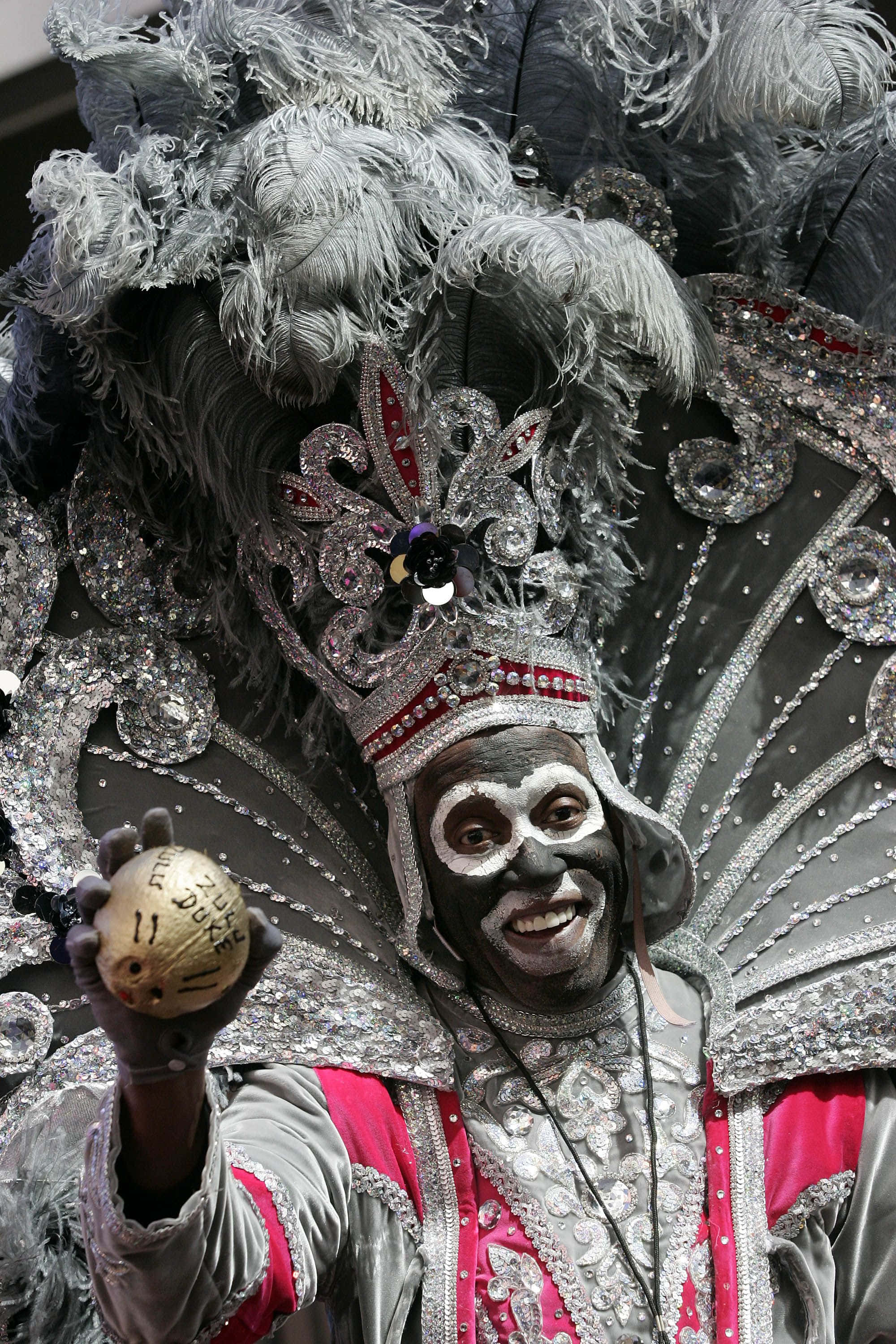 Mardi Gras 桌布 壁紙 — 一位快樂的表演者穿著華麗的銀色和粉紅色嘉年華服裝，戴著羽毛頭飾，手持金色球。