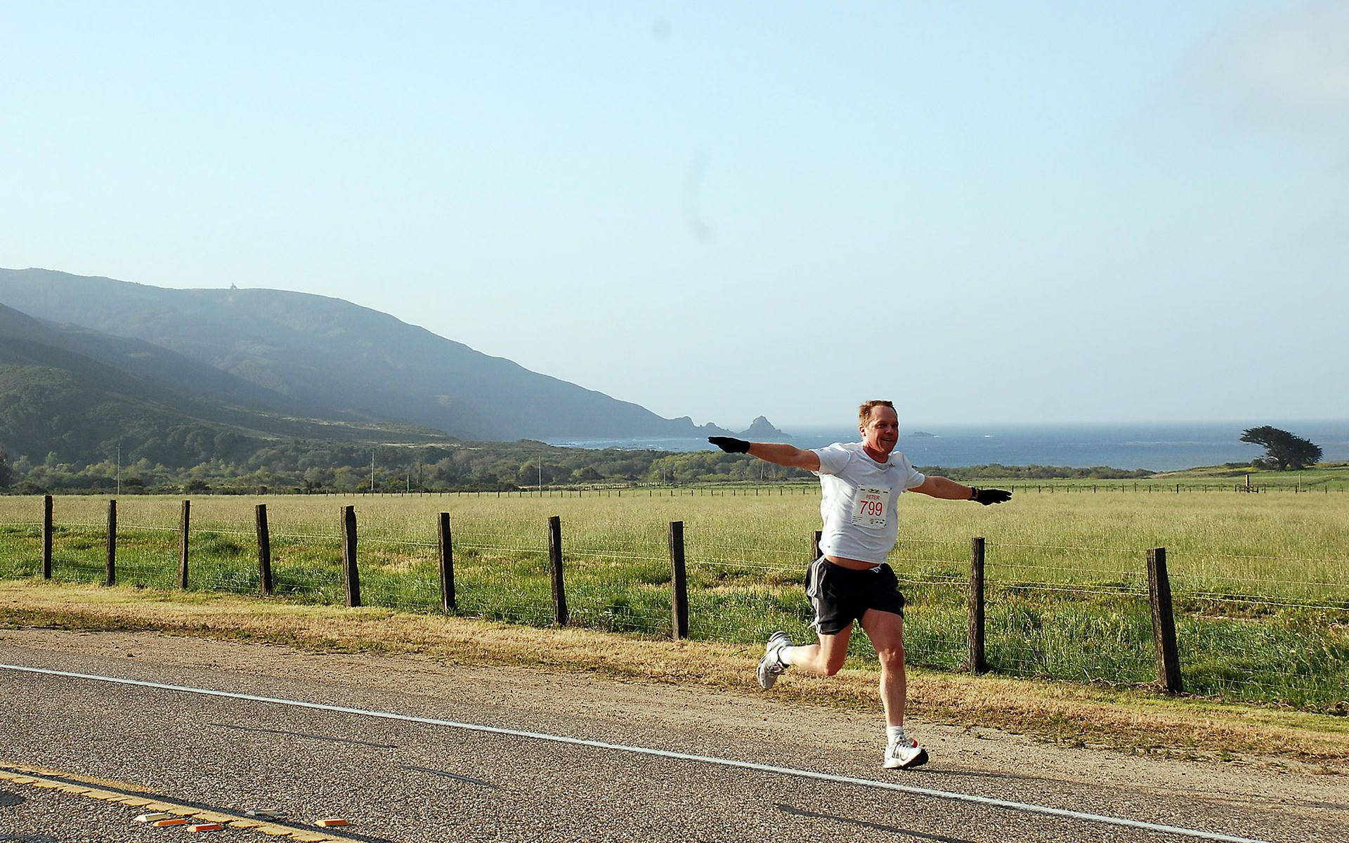 Marathon 桌布 壁紙 — 一位快樂的跑者在沿海道路上張開雙臂，對著起伏的綠色山丘和海洋地平線慶祝。