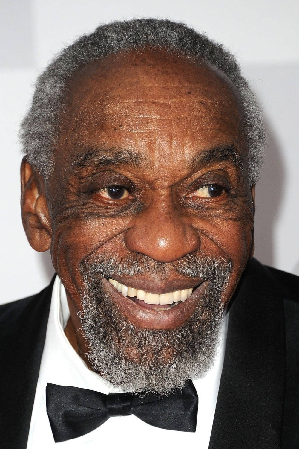 actor 桌布 壁紙 — 一位快樂的長者，留著灰色鬍鬚，帶著溫暖的笑容，身穿黑色禮服和領結，背景為純白色。