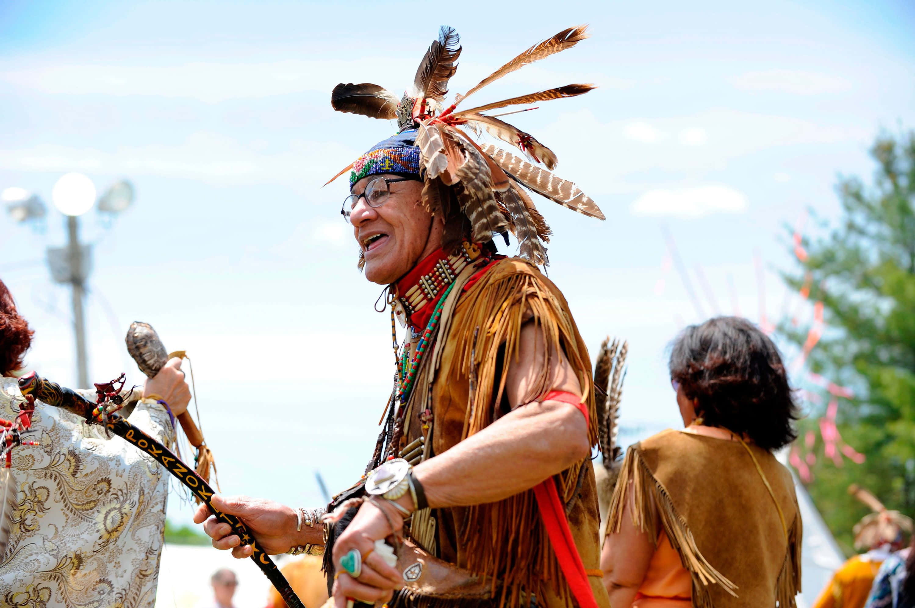 Native American 桌布 壁紙 — 一位快樂的長者穿著傳統美洲原住民服飾，頭戴羽毛頭飾，在戶外節慶中微笑著手持儀式用杖。
