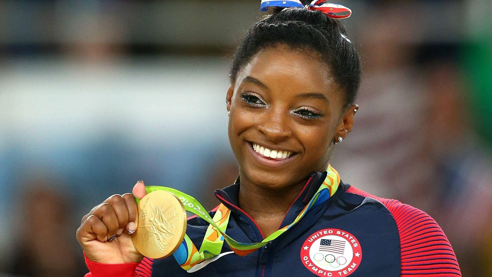 Simone Biles 桌布 壁紙 — 一位快樂的體操運動員在充滿活力的場館中自豪地高舉她的奧運金牌，笑容燦爛。