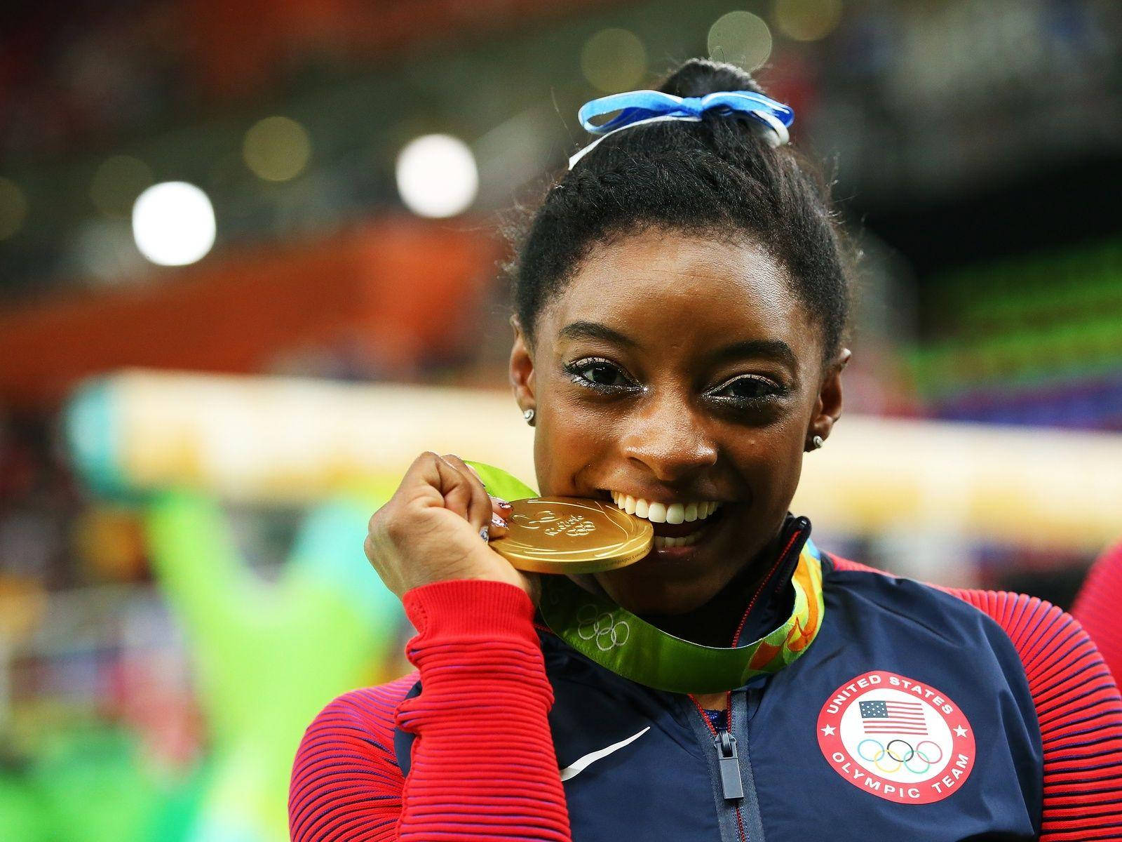 Gold Medal 桌布 壁紙 — 一位快樂的體操運動員在體育場內穿著美國隊外套，咬著她的奧運金牌。