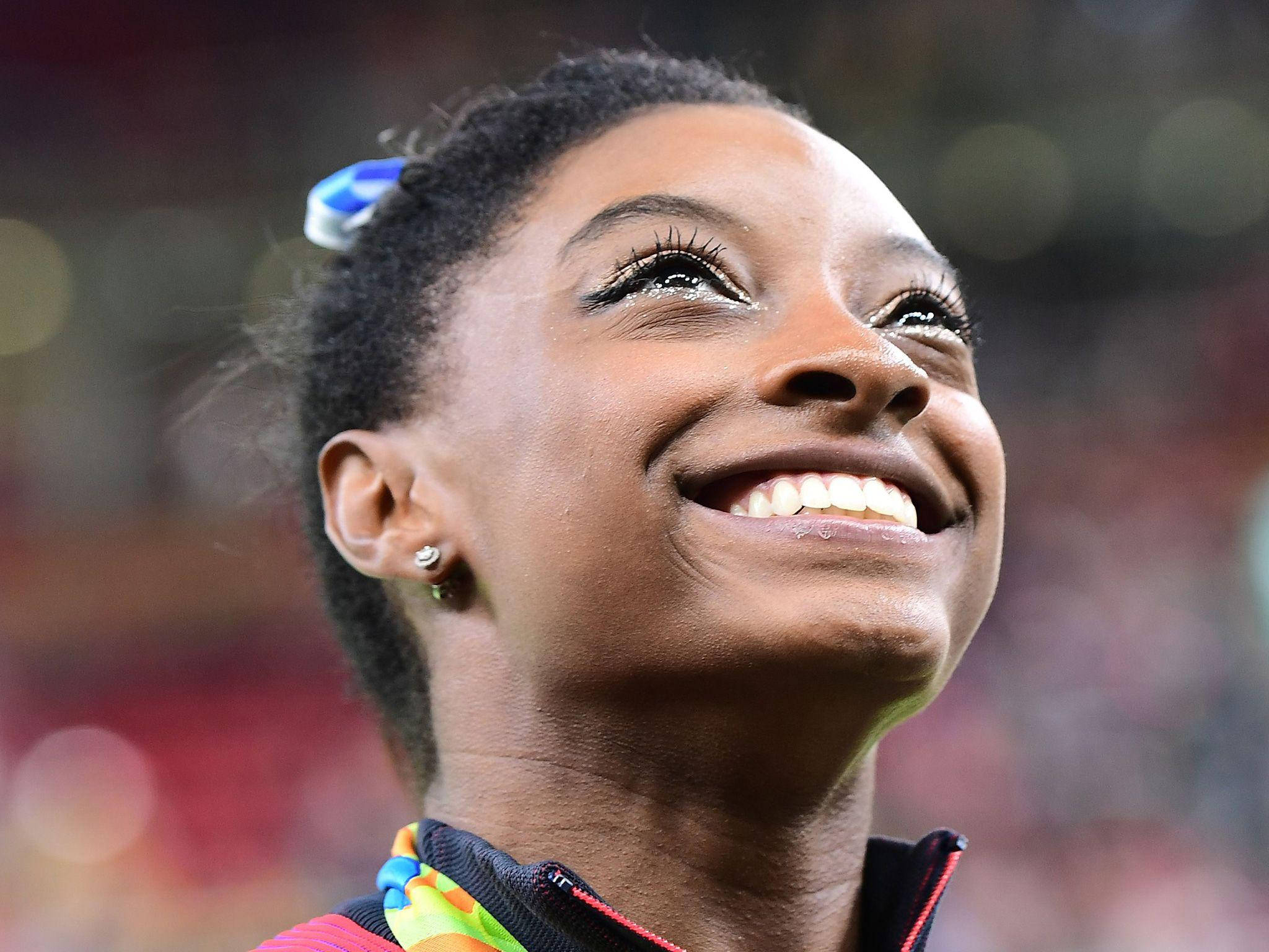 Simone Biles 桌布 壁紙 — 一位快樂的體操運動員在體育場環境中，脖子上掛著彩色獎牌，自豪地閃耀。