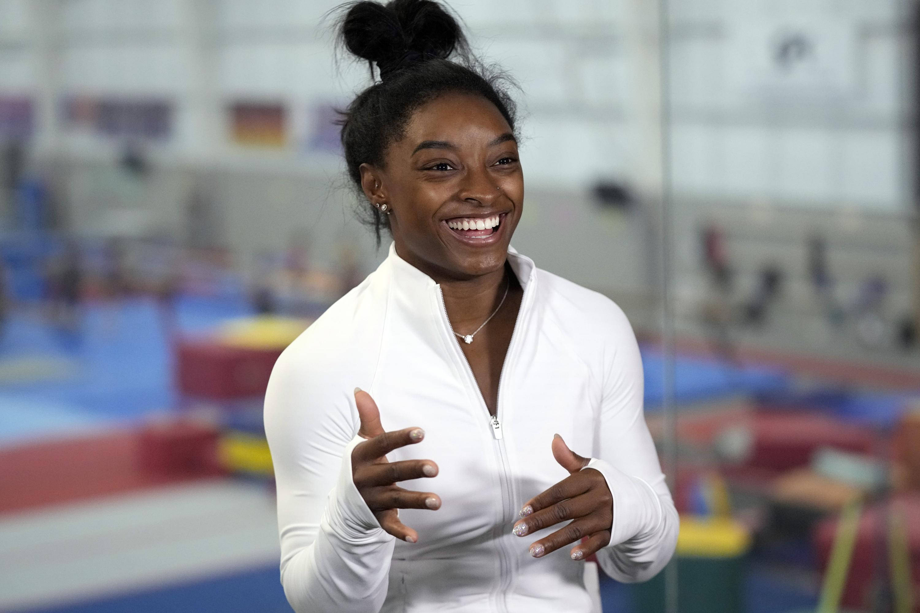 simon biles 桌布 壁紙 — 一位快樂的體操運動員穿著白色拉鍊外套，在充滿彩色設備的體育館中張開雙手燦爛微笑。