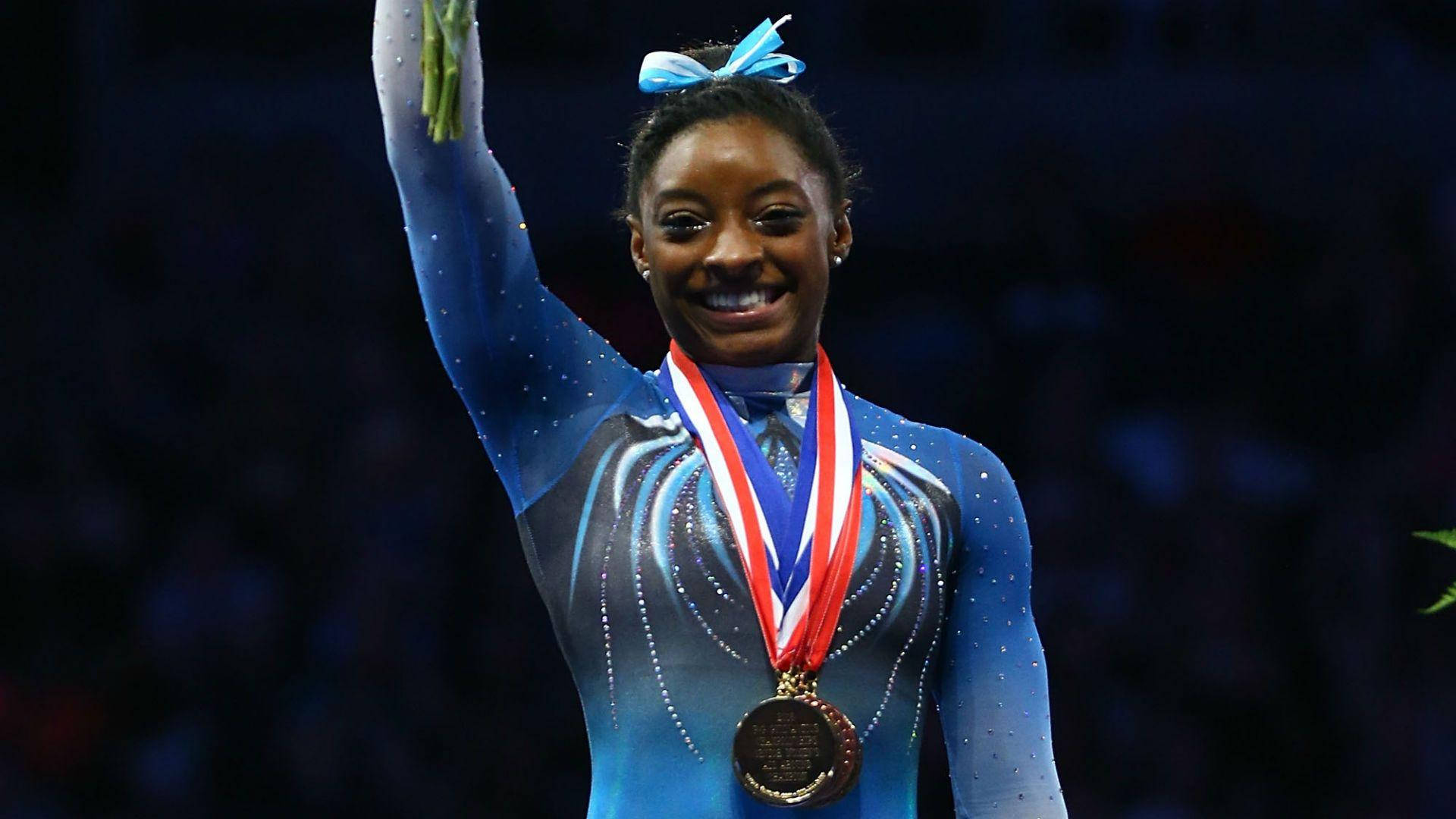 Simone Biles 桌布 壁紙 — 一位快樂的體操運動員穿著藍色緊身衣，揮舞著花束，脖子上自豪地掛著金牌。