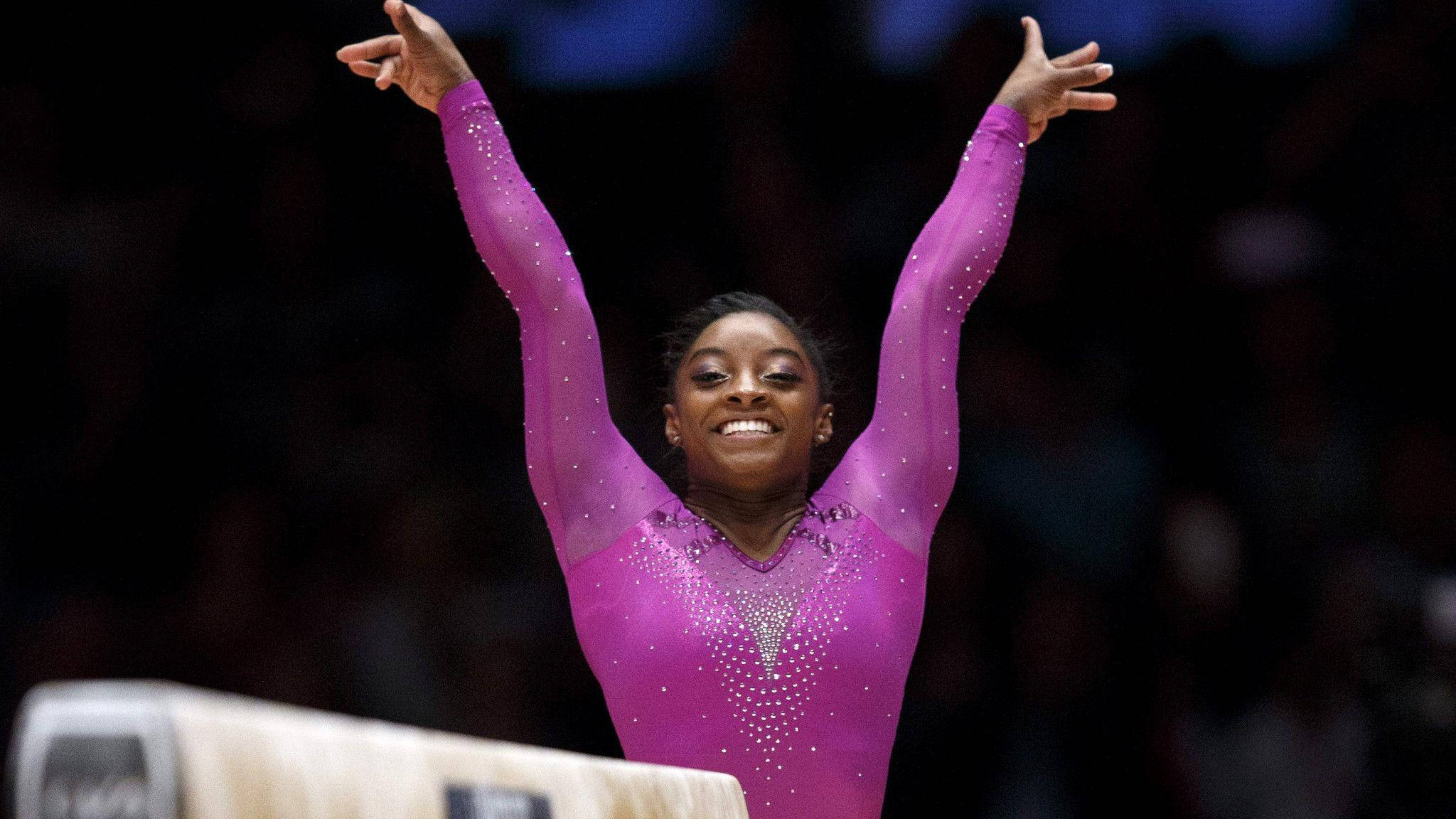 Simone Biles 桌布 壁紙 — 一位快樂的體操運動員穿著閃亮的紫色緊身衣，在平衡木上完成動作後勝利地舉起雙臂。