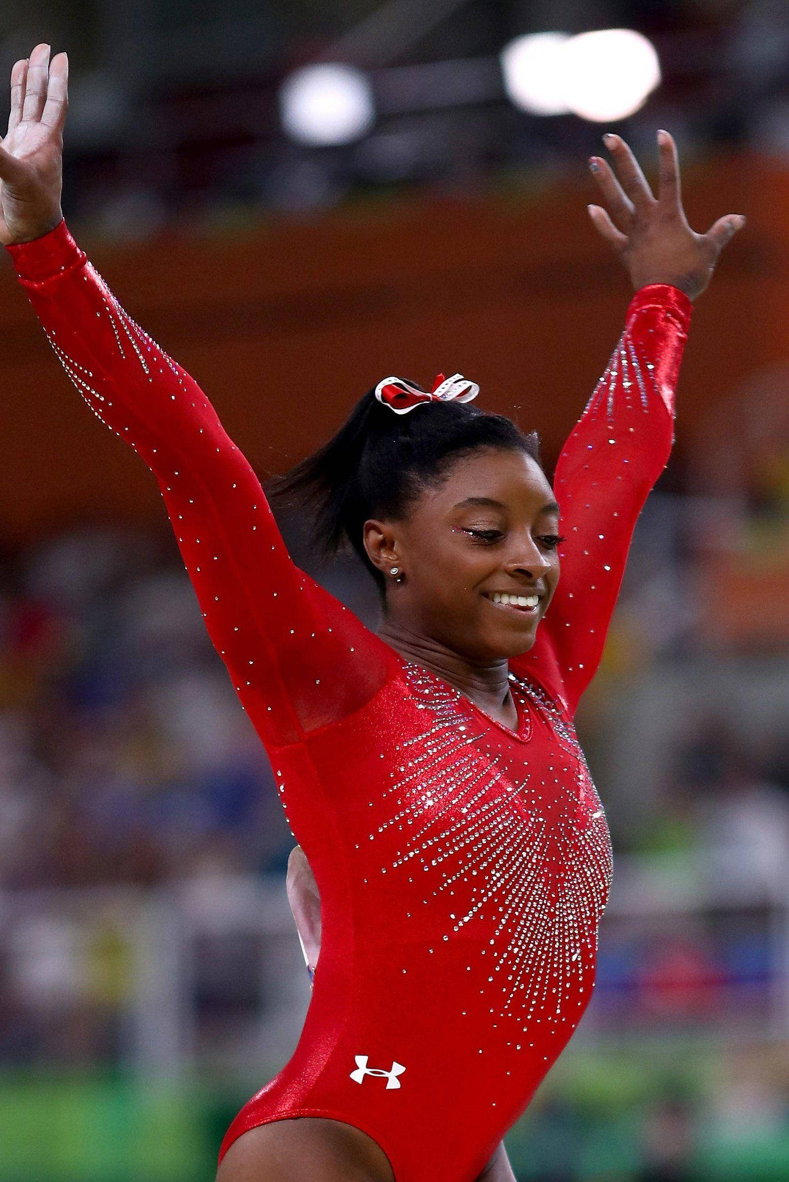 Gymnastics Free 桌布 壁紙 — 一位快樂的體操運動員穿著閃閃發光的紅色緊身衣，在明亮的場館燈光下，於比賽套路中高舉雙臂慶祝勝利。
