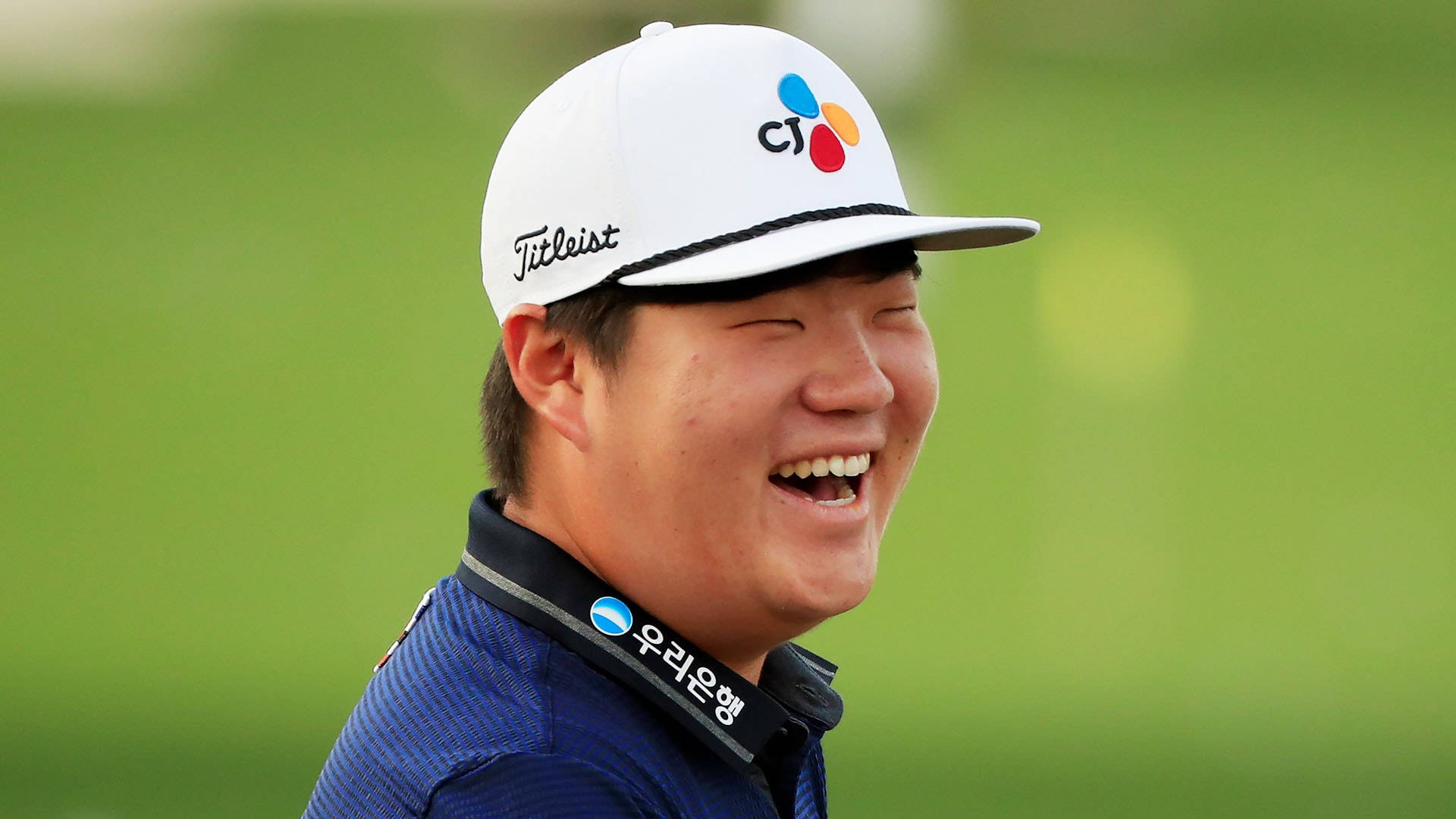 運動 桌布 壁紙 — 一位快樂的高爾夫球手戴著白色Titleist帽子和藍色Polo衫，在模糊的綠色背景下開懷大笑。