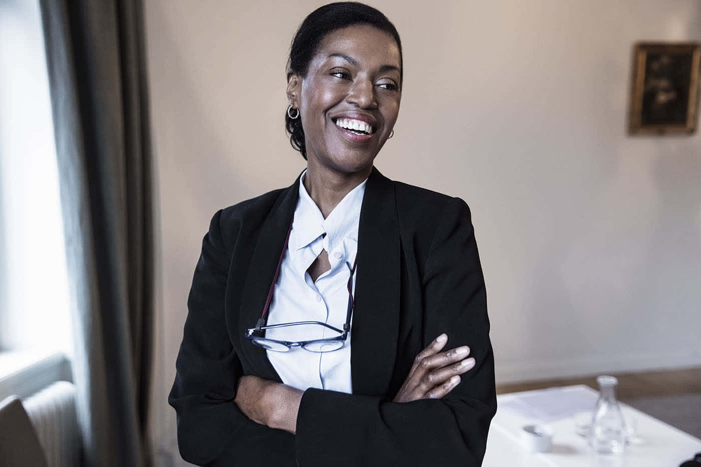 Lawyer 桌布 壁紙 — 一位快樂的黑人女性，身穿黑色西裝外套和白色襯衫，在室內窗邊交叉雙臂，溫暖地微笑。