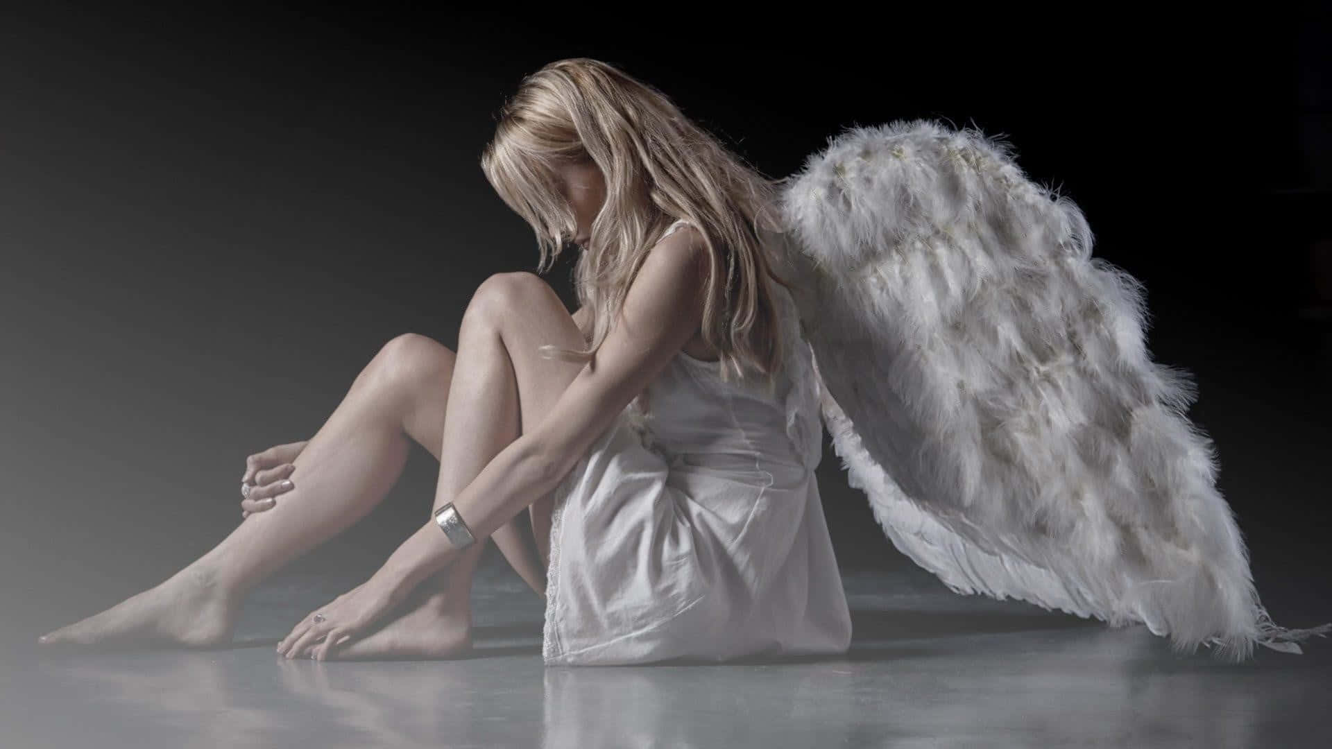 Beautiful Angel 桌布 壁紙 — 一位悲傷的天使，長著白色羽毛翅膀，蜷縮在昏暗工作室的反光地板上，低垂著頭，陷入無聲的絕望。