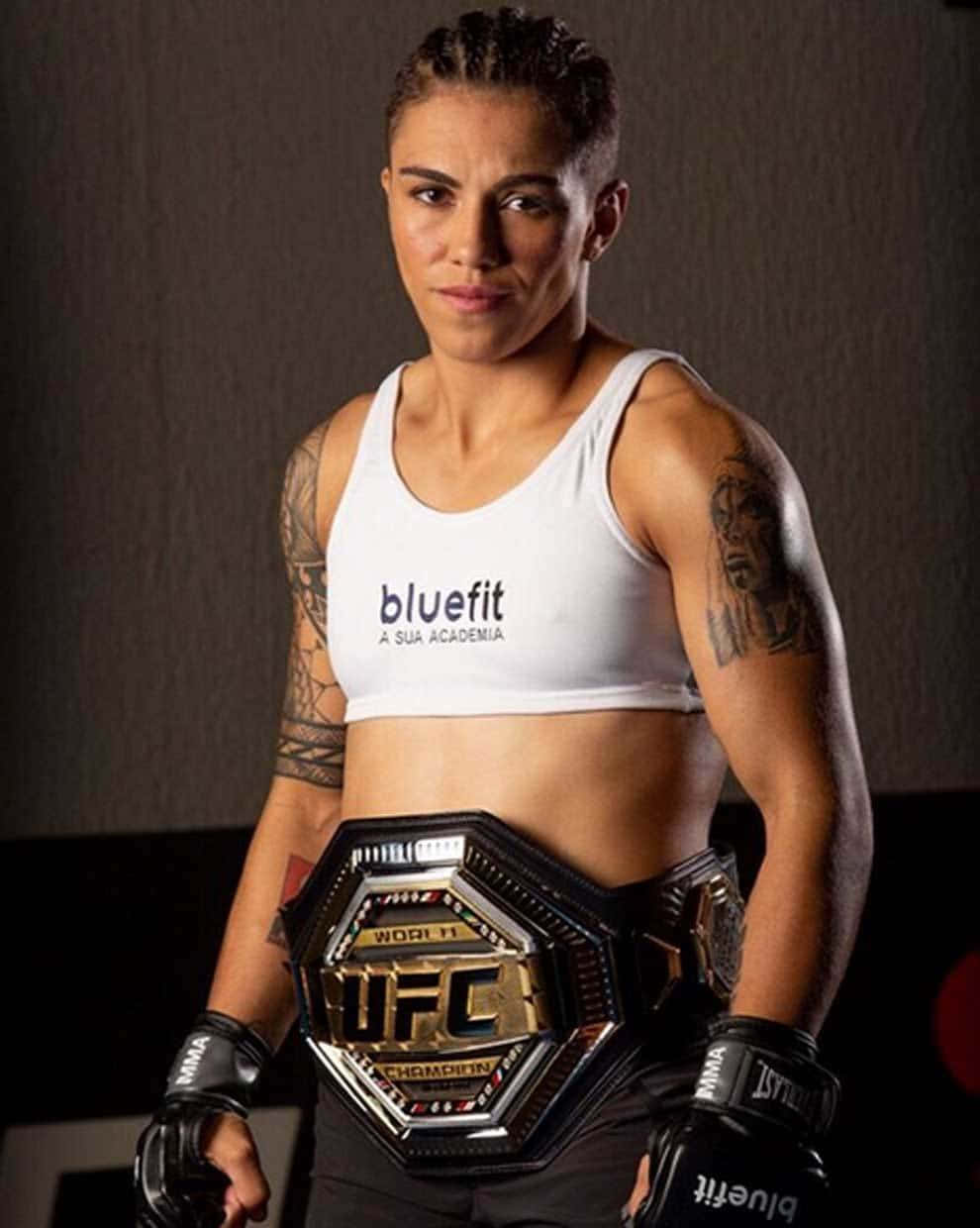MMA 桌布 壁紙 — 一位意志堅定的女性MMA拳手，身穿白色背心和黑色手套，在深色攝影棚背景下自豪地高舉UFC冠軍腰帶。