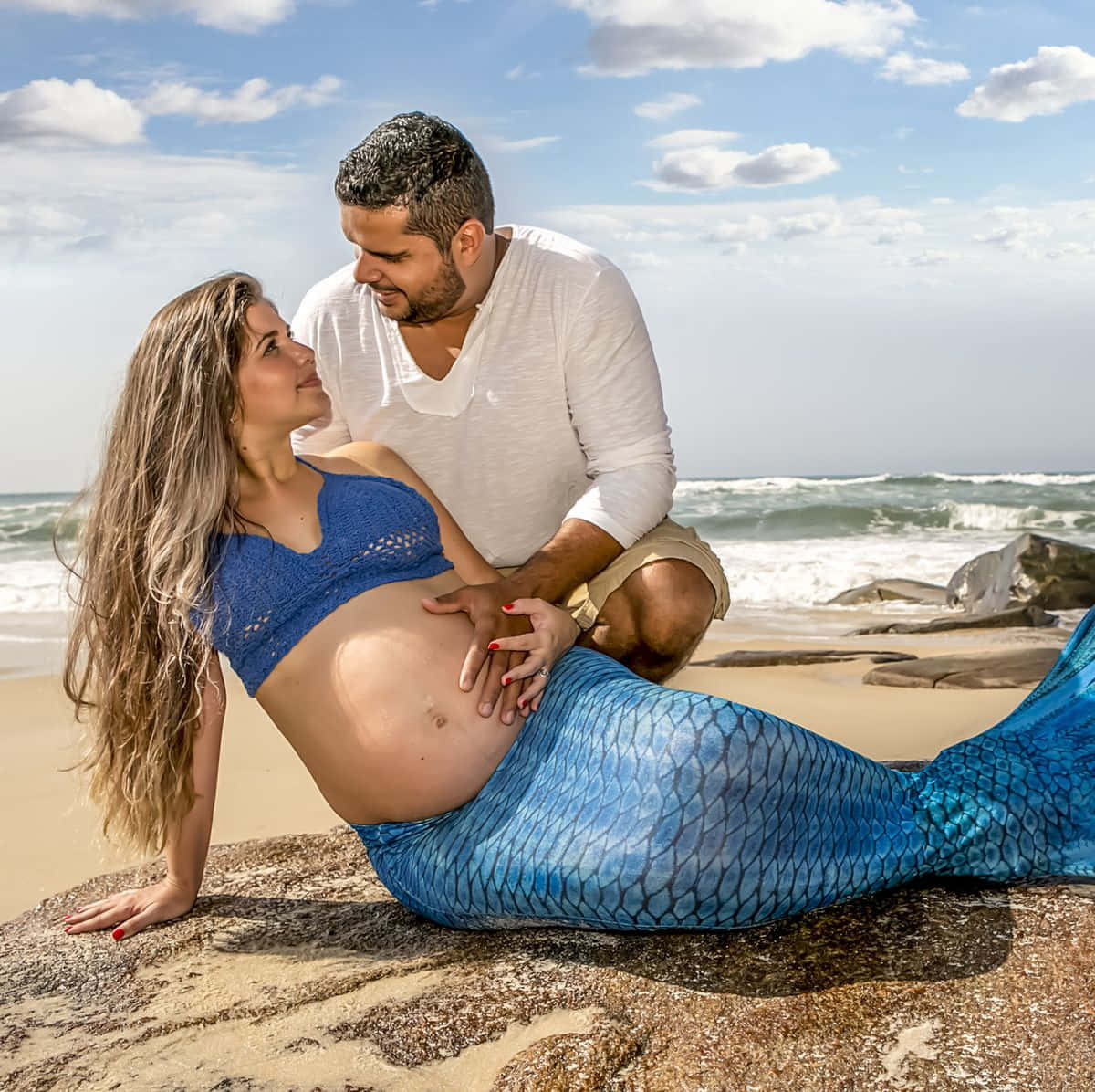 Mermaids 桌布 壁紙 — 一位懷孕的女性穿著藍色美人魚尾巴在岩石上休息，她的伴侶在陽光明媚的海灘上輕柔地觸摸她的肚子。