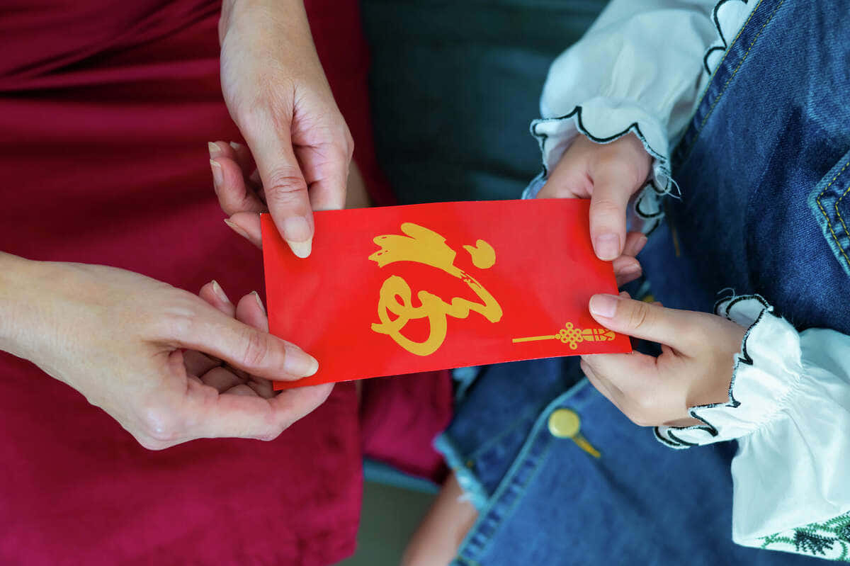 Red Envelope 桌布 壁紙 — 一位成年人將一個帶有金色中文字的紅色紅包遞給穿著牛仔褲和白色襯衫的小孩。