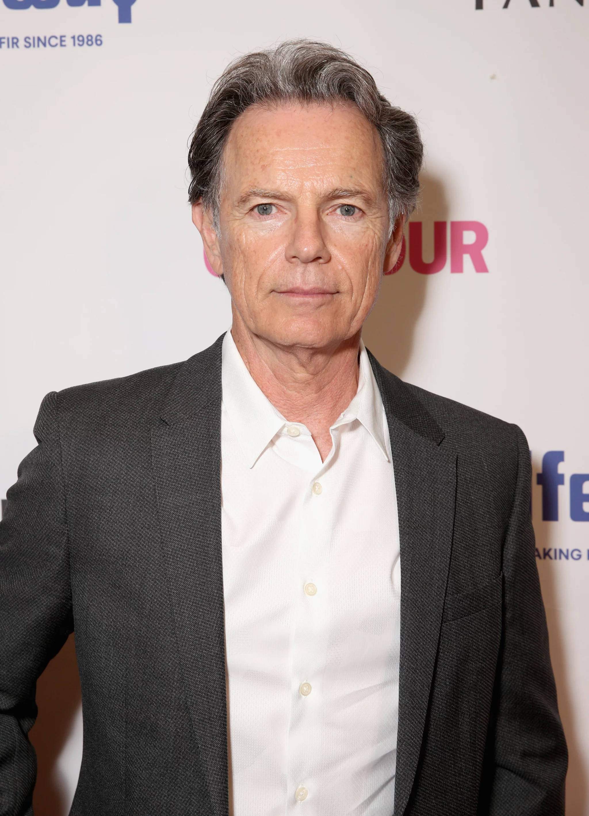 actor 桌布 壁紙 — 一位成熟男士，灰髮藍眼，身穿深色西裝和白襯衫，背景為品牌活動場景。