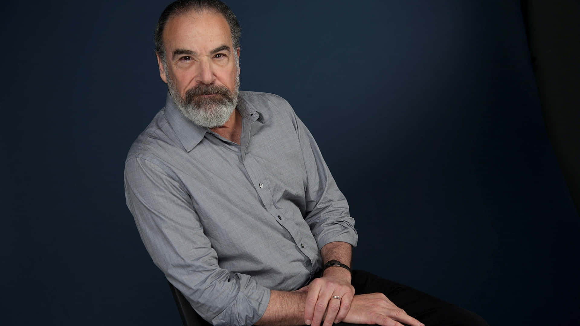 Mandy Patinkin 桌布 壁紙 — 一位成熟男士留著灰色鬍鬚，自信地坐在椅子上，背景是深藍色攝影棚，身穿灰色鈕扣襯衫。
