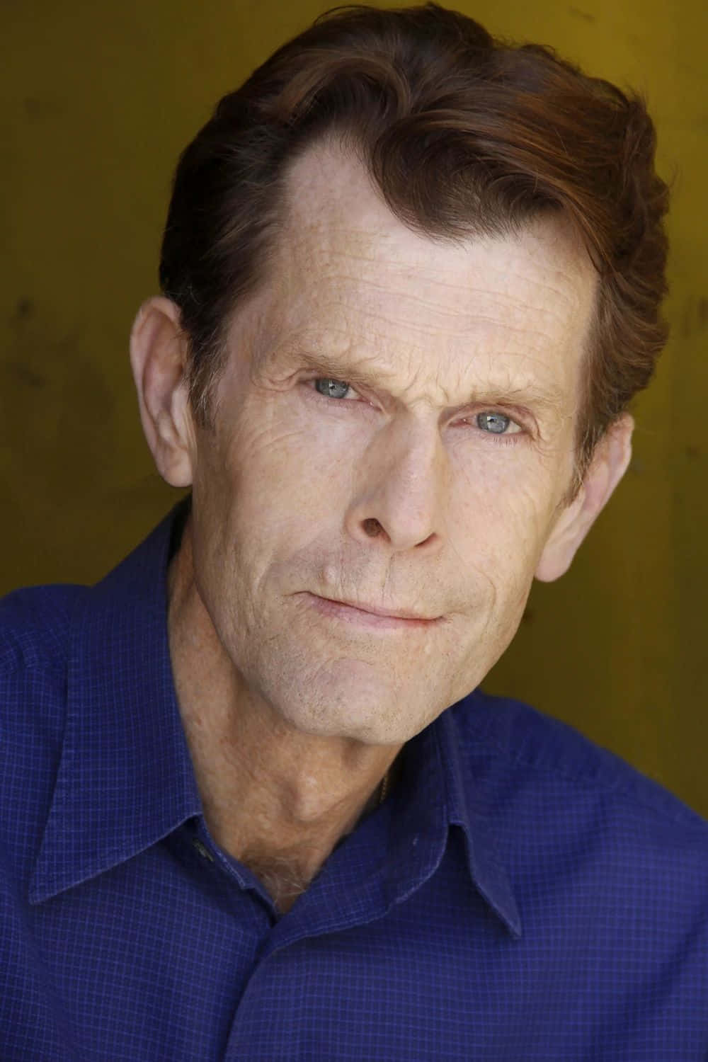 Kevin Conroy 桌布 壁紙 — 一位成熟男士的特寫肖像，擁有強烈的藍色眼睛和波浪形棕色頭髮，穿著藍色格紋襯衫，背景是溫暖的黃色。