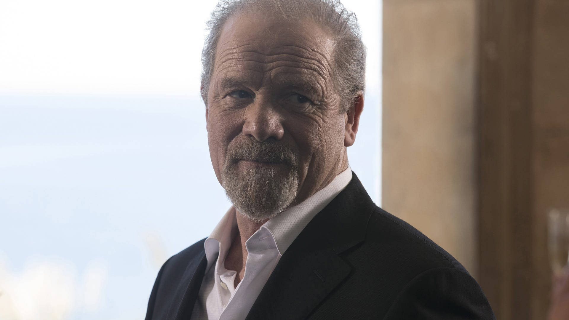 HBO 桌布 壁紙 — 一位成熟男士的近距离肖像，灰发胡须，身穿黑色西装和白色衬衫，直视镜头，带着微妙的微笑。