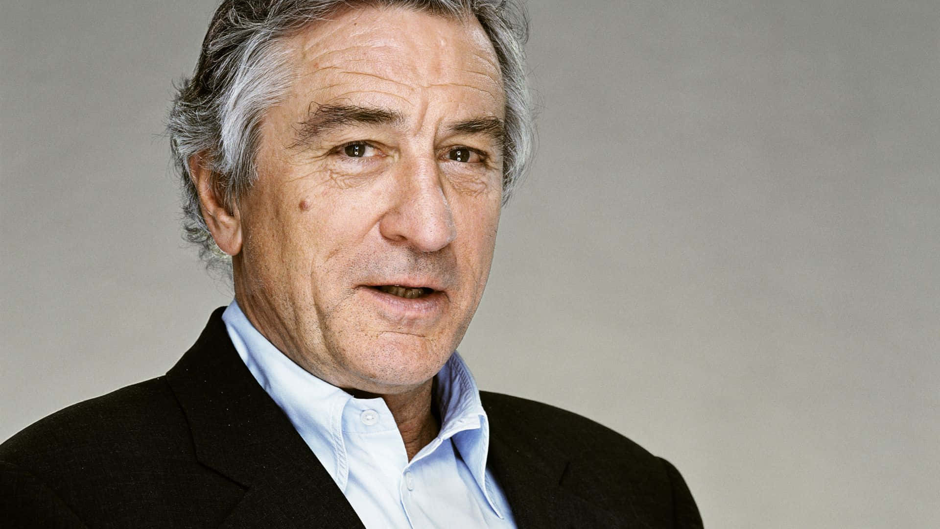 actor 桌布 壁紙 — 一位成熟男士的頭像特寫，灰髮，身穿黑色西裝外套內搭淺藍色襯衫，背景為中性灰色。