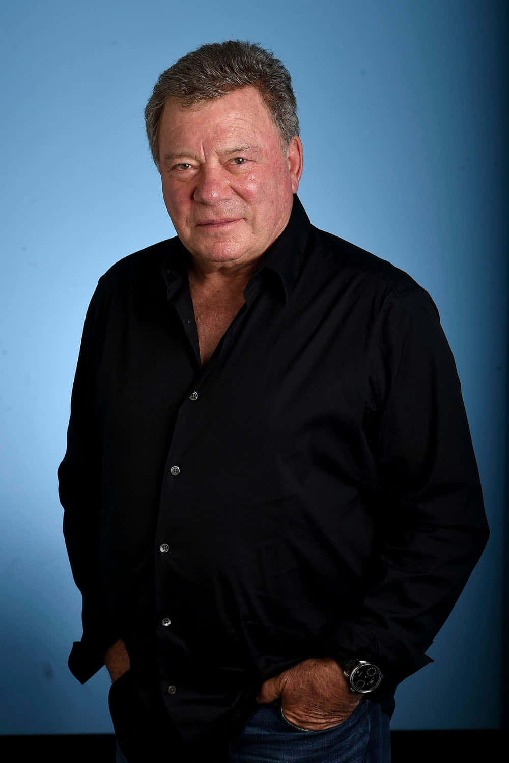 william shatner 桌布 壁紙 — 一位成熟男士穿著黑色襯衫，雙手插袋，自信地站在純藍色攝影棚背景前。