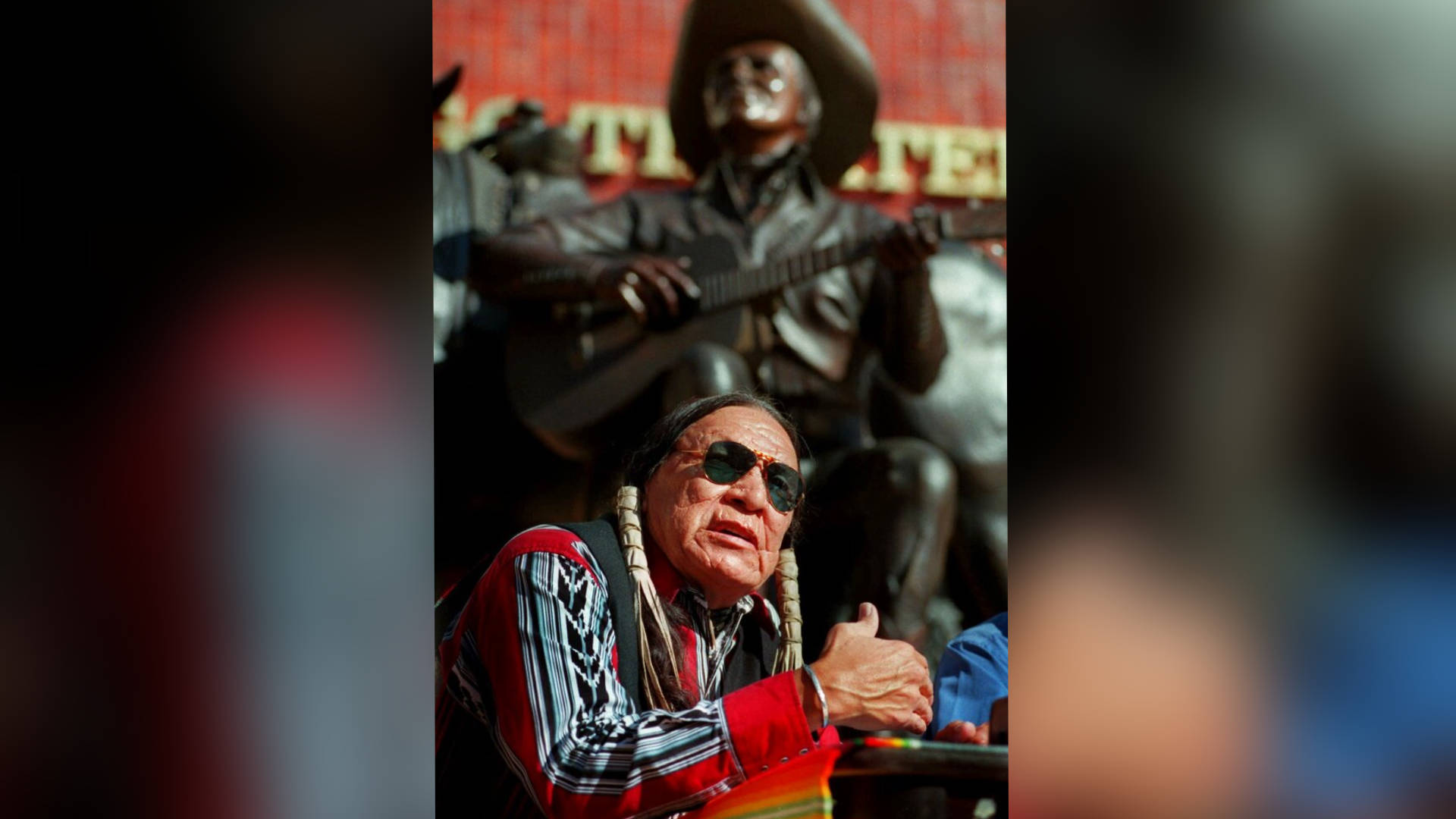Native American 桌布 壁紙 — 一位戴太陽眼鏡和圖案襯衫的長者在吉他手銅像前豎起大拇指。