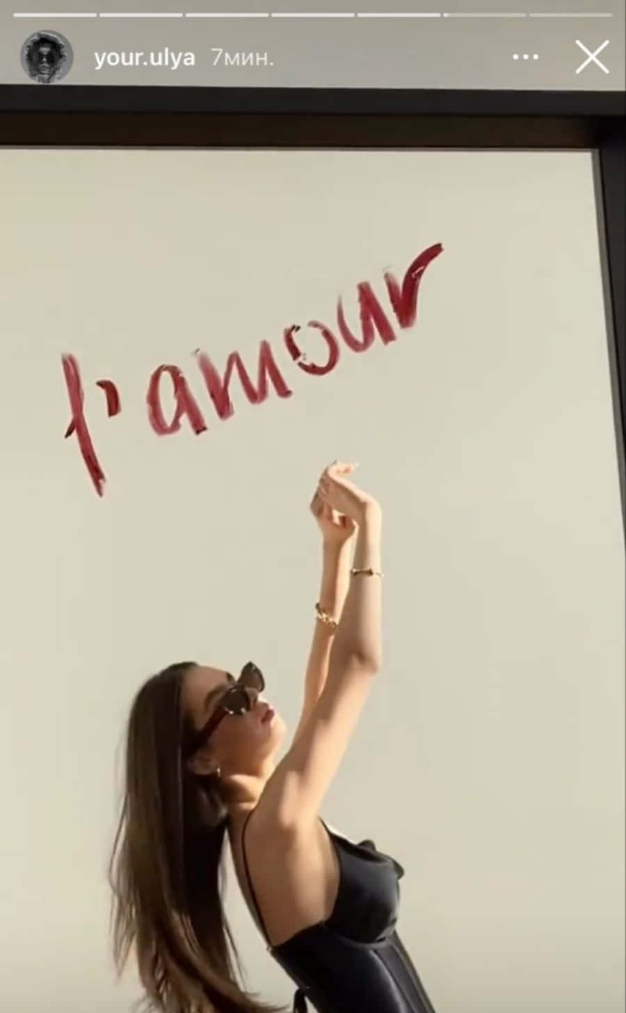 Instagram Story 桌布 壁紙 — 一位戴太陽眼鏡的女性向上伸手，觸向白色牆上以紅色草書寫成的「l'amour」一詞。