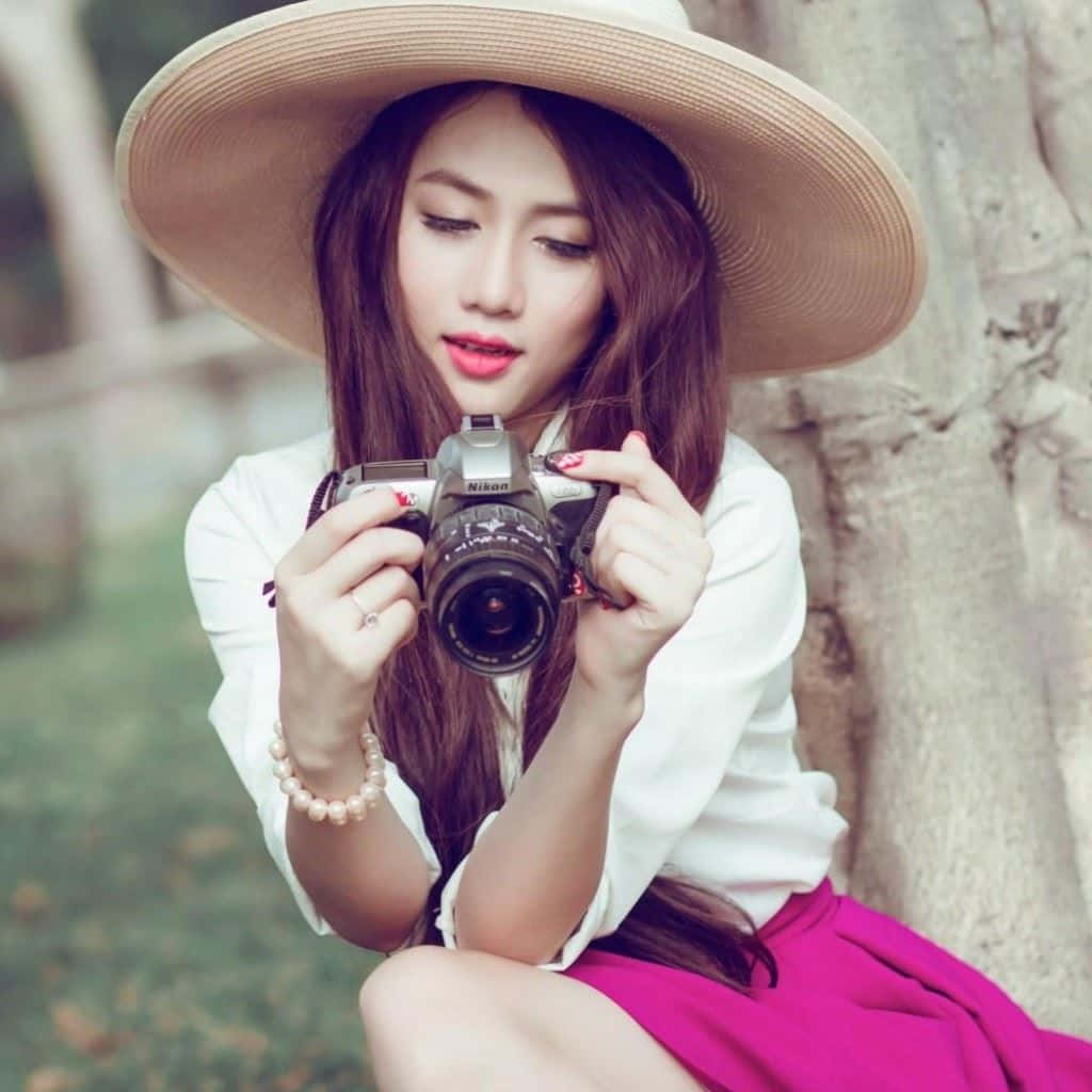 美女 桌布 壁紙 — 一位戴寬邊帽、穿白色襯衫的年輕女子坐在花園中一棵樹旁，手持一部復古相機，背景柔和。