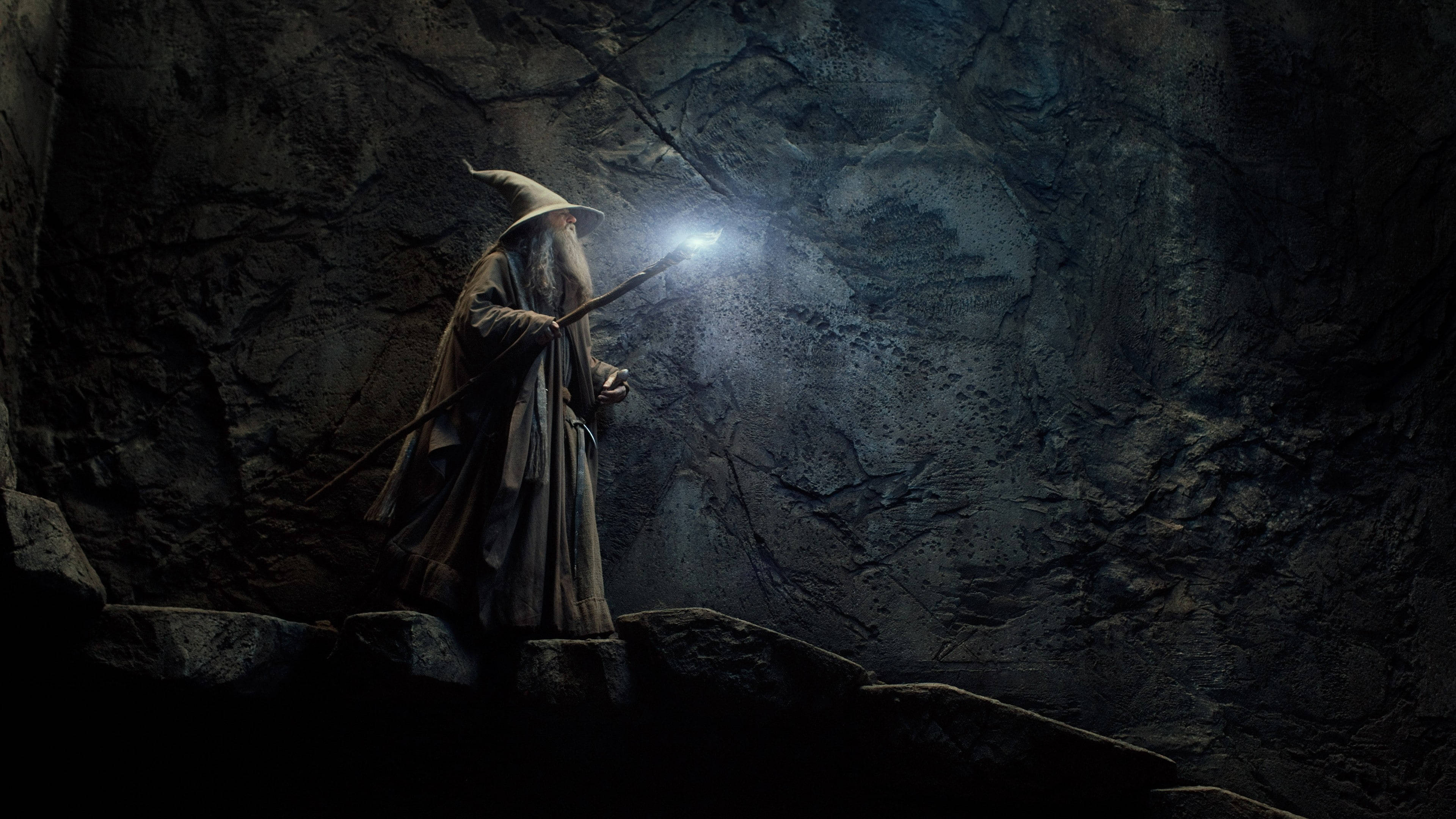Lord Of The Rings Landscape 桌布 壁紙 — 一位戴尖帽的巫師手持發光法杖，照亮黑暗的岩石洞穴。