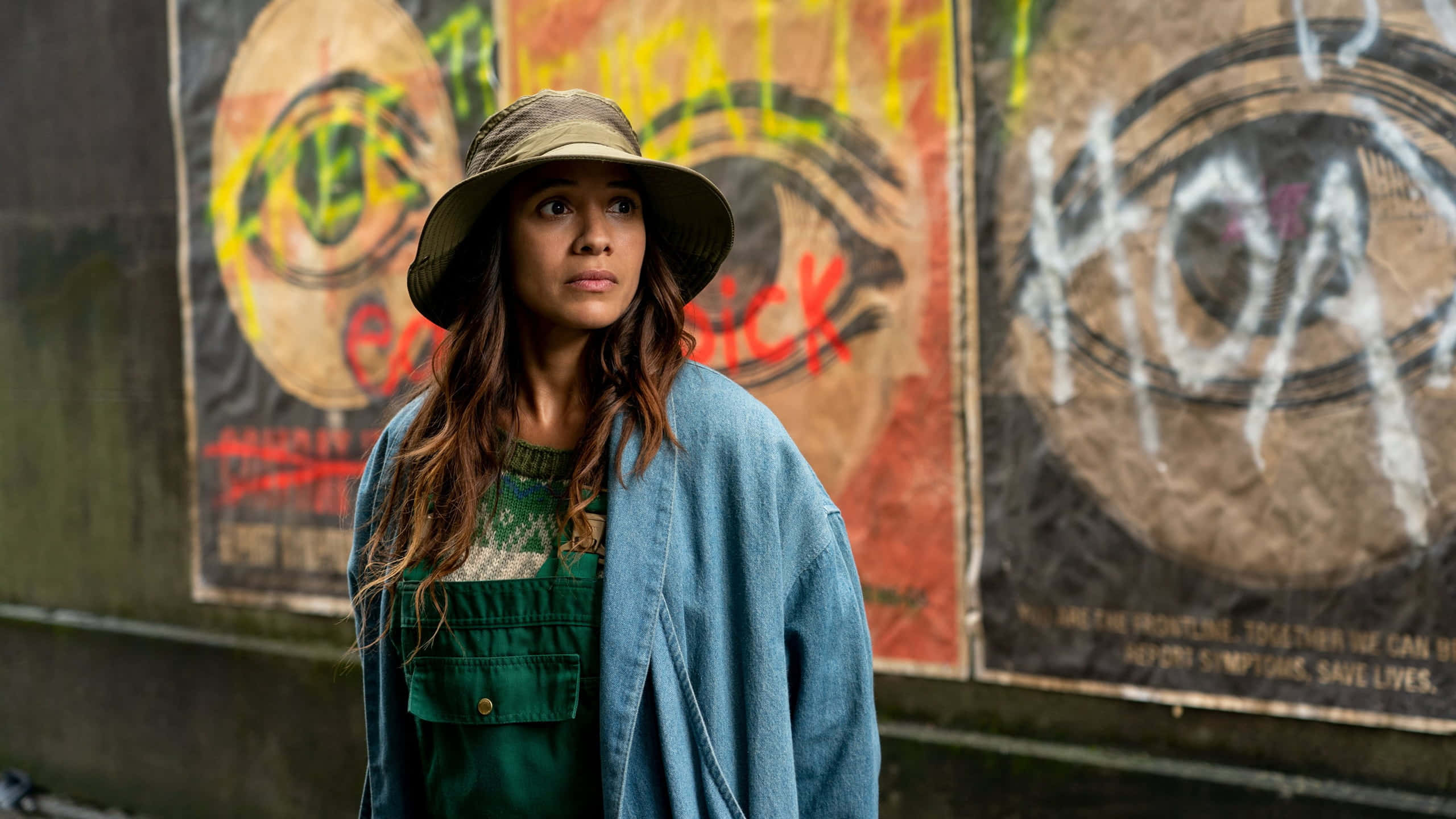 Dania Ramirez 桌布 壁紙 — 一位戴帽子和牛仔外套的女性站在佈滿塗鴉的海報前，海報上有大膽的眼睛和抗議標語。