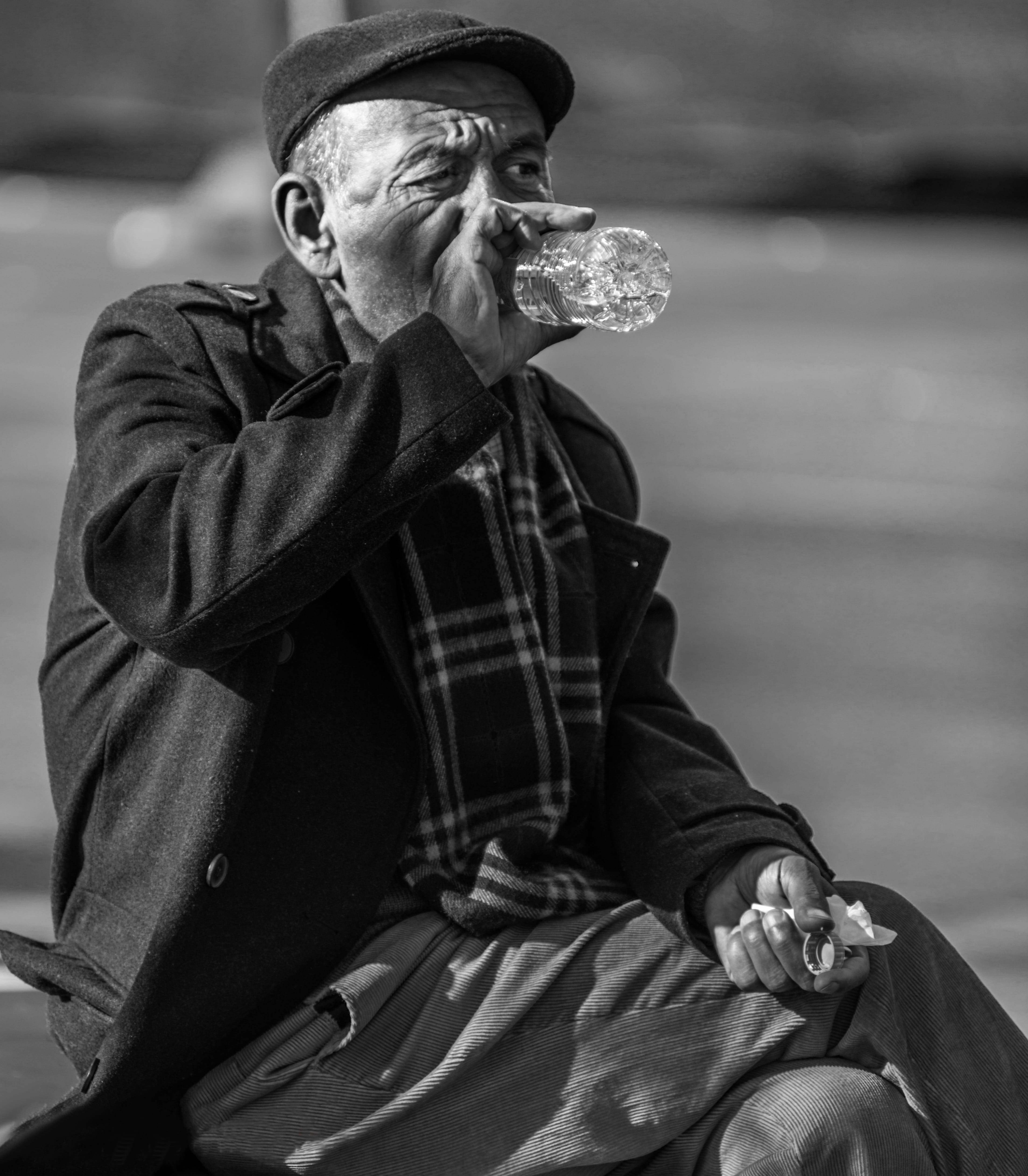 Drinking 桌布 壁紙 — 一位戴帽子和穿外套的長者，在戶外黑白環境中坐著，從塑膠瓶喝水。