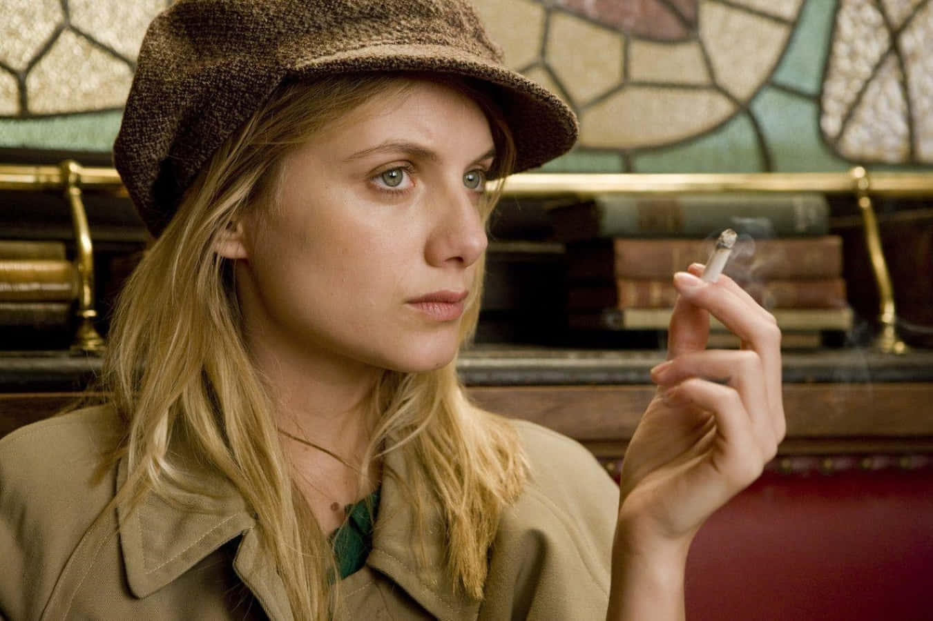 Mélanie Laurent 桌布 壁紙 — 一位戴棕色帽子的女士在一個帶有彩色玻璃和書架的復古房間裡拿著一支香煙。