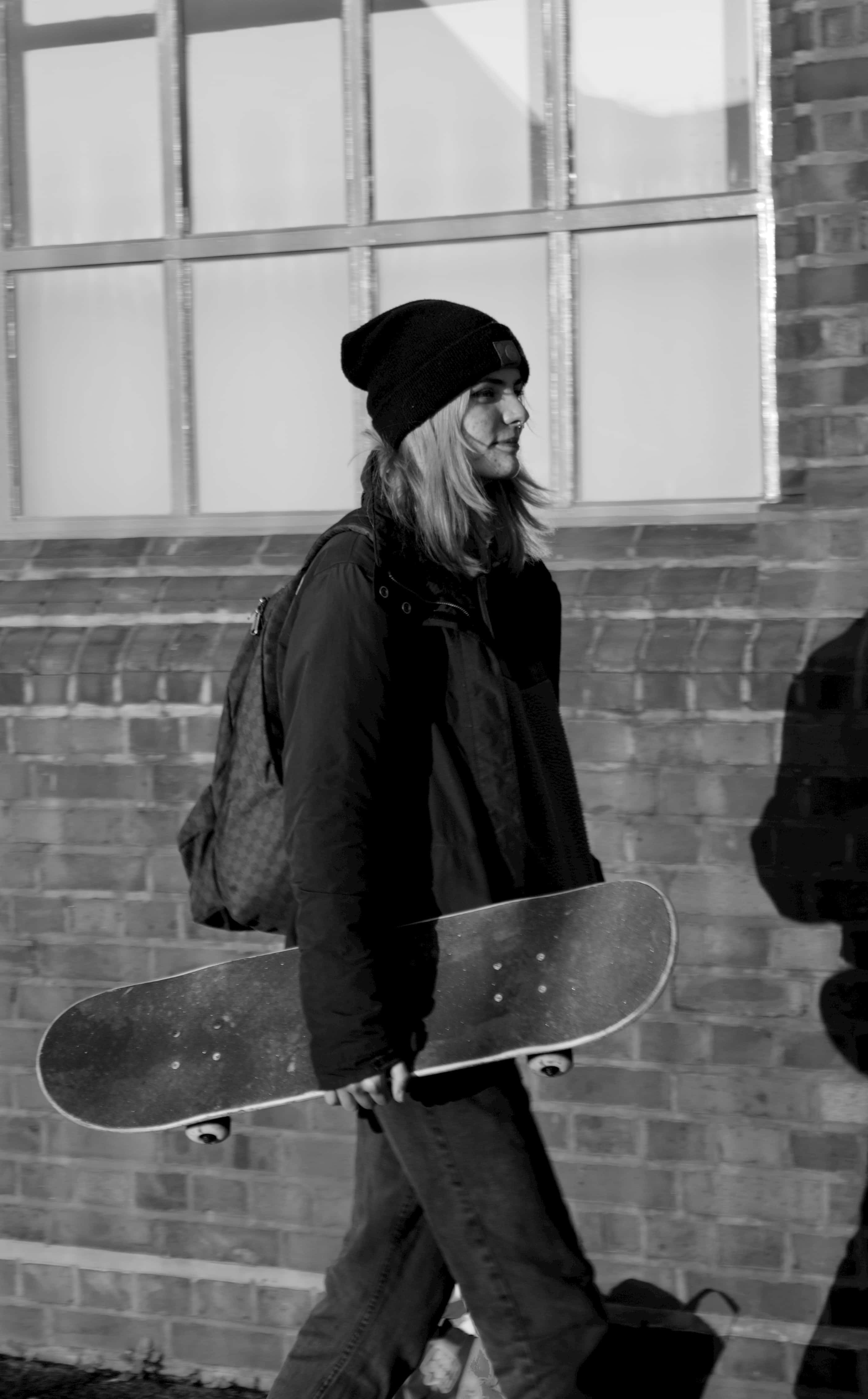 Skater Girl Aesthetic 桌布 壁紙 — 一位戴毛帽和穿外套的年輕人拿著滑板走過磚牆，黑白風格。