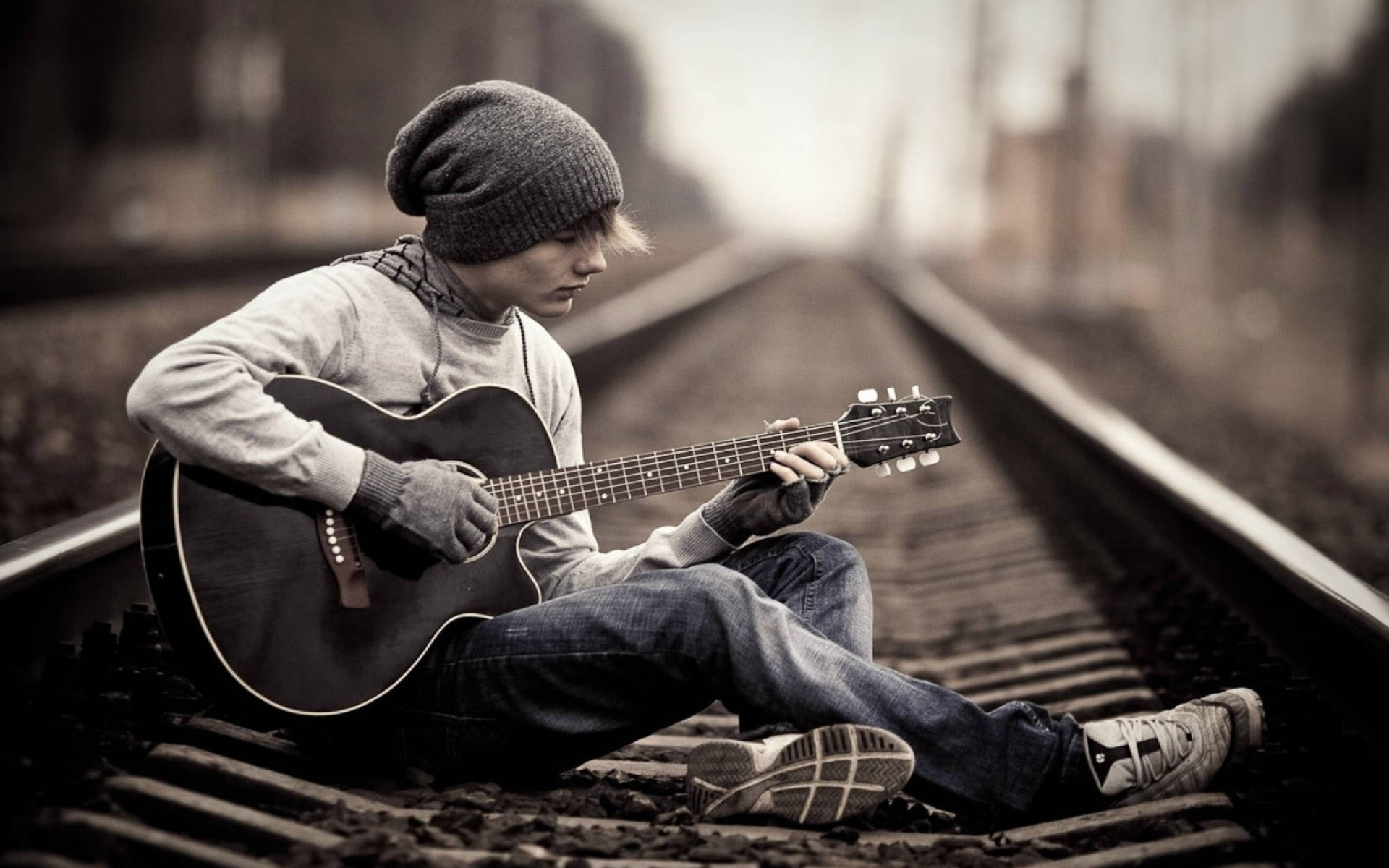 Alone 桌布 壁紙 — 一位戴毛帽的年輕音樂家盤腿坐在鐵軌上，在朦朧的天空下彈奏木吉他。