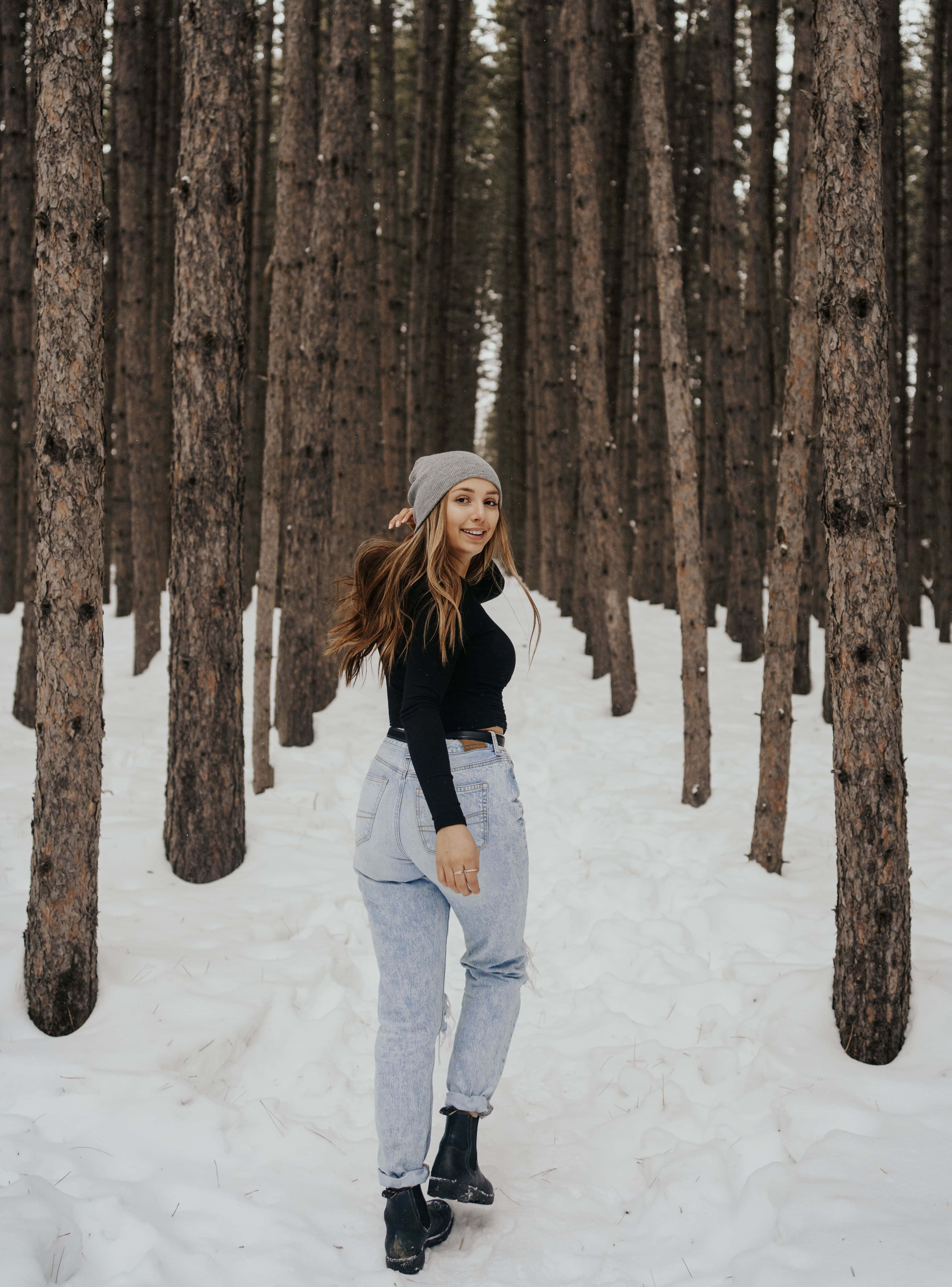Winter Photography 桌布 壁紙 — 一位戴灰色毛帽、穿淺色牛仔褲的微笑女士站在被新雪覆蓋的高大松樹之間。