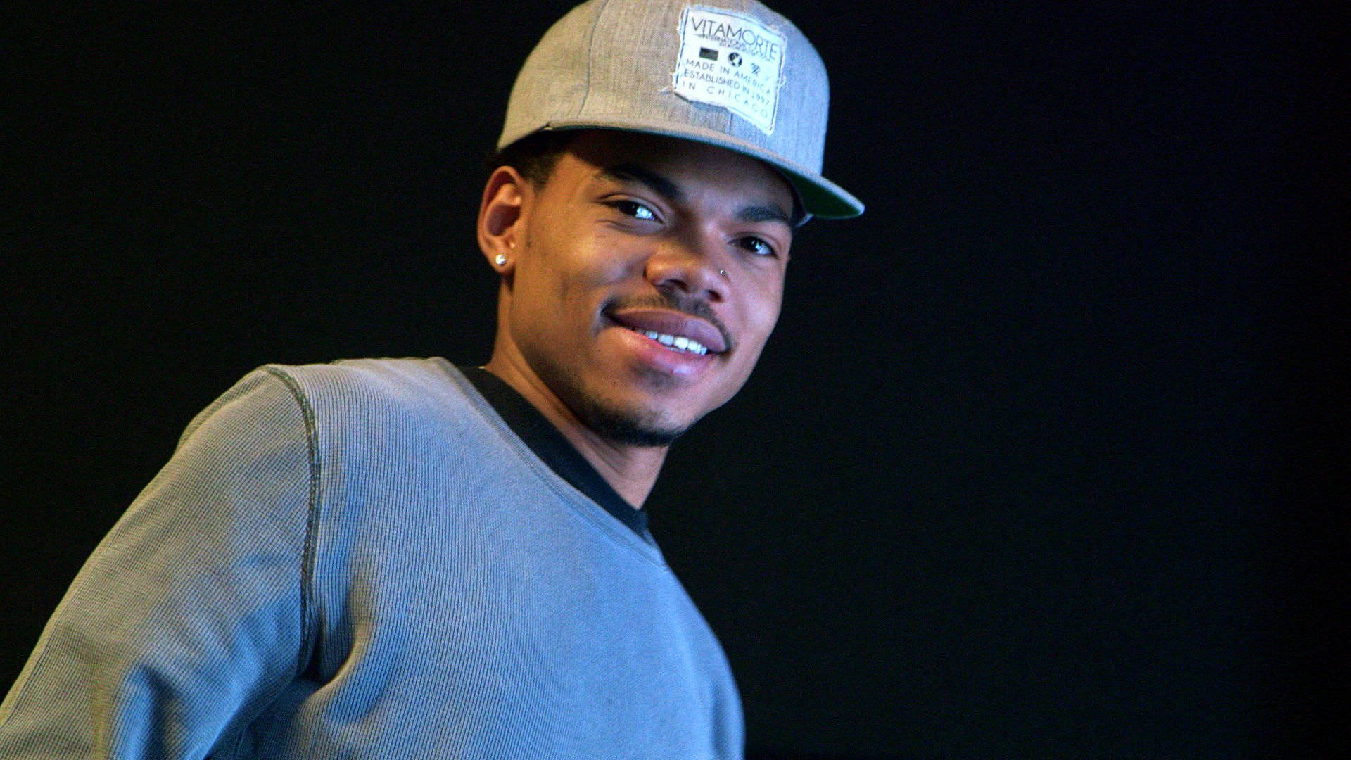 chance 桌布 壁紙 — 一位戴灰色鴨舌帽和穿毛衣的年輕男子在深色背景前自信地微笑。