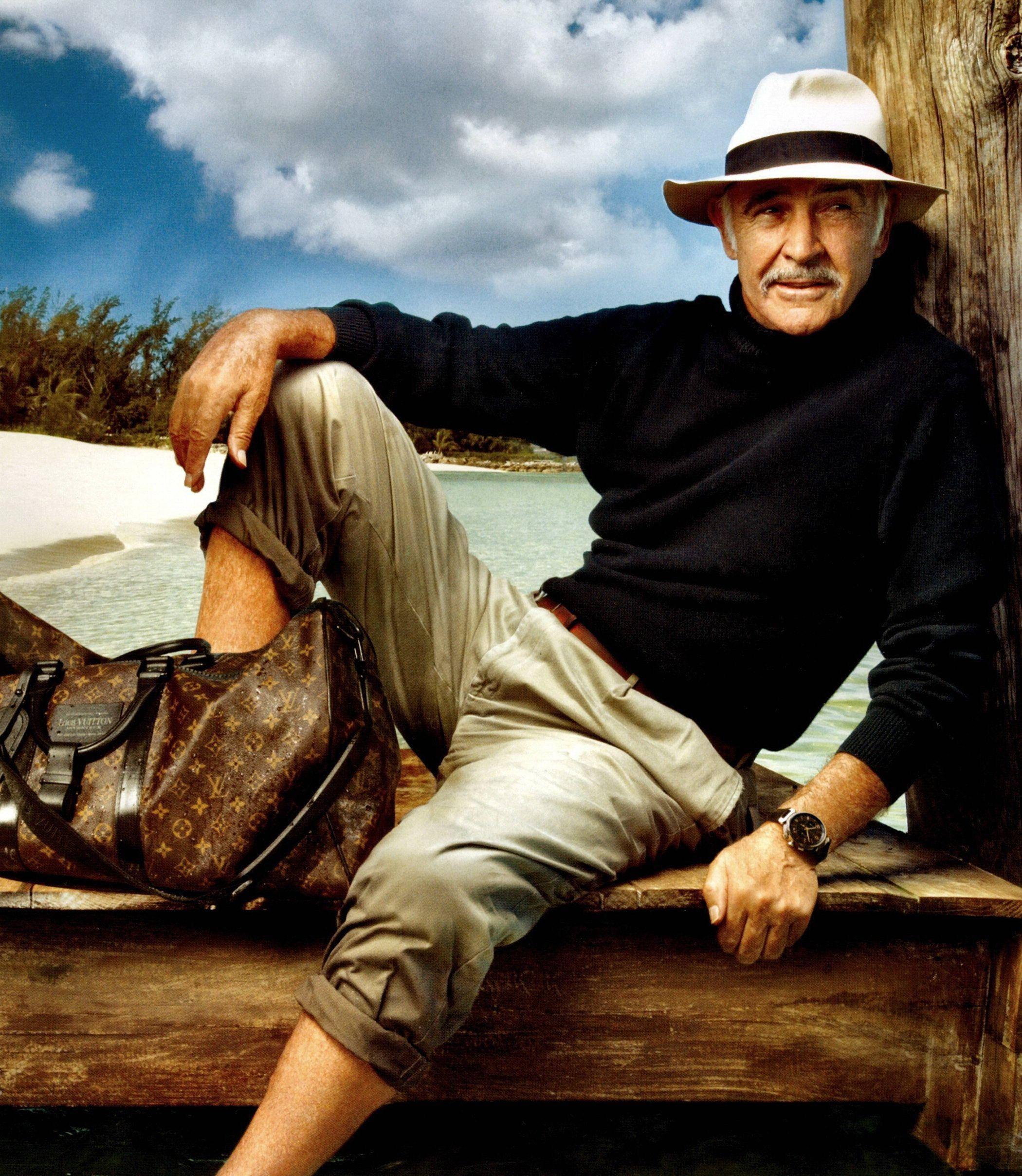 Sean Connery 桌布 壁紙 — 一位戴白帽、穿黑色毛衣的年長男子在木碼頭上放鬆，旁邊是碧綠的海水和白色的沙灘，天空部分多雲。