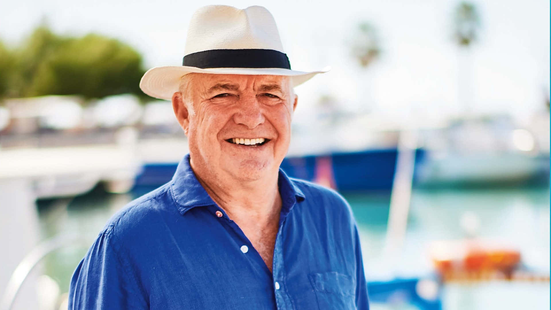 Beach Hat 桌布 壁紙 — 一位戴白帽穿藍衫的快樂長者，在陽光明媚的碼頭微笑，背景有船和棕櫚樹，呈現柔和焦點。