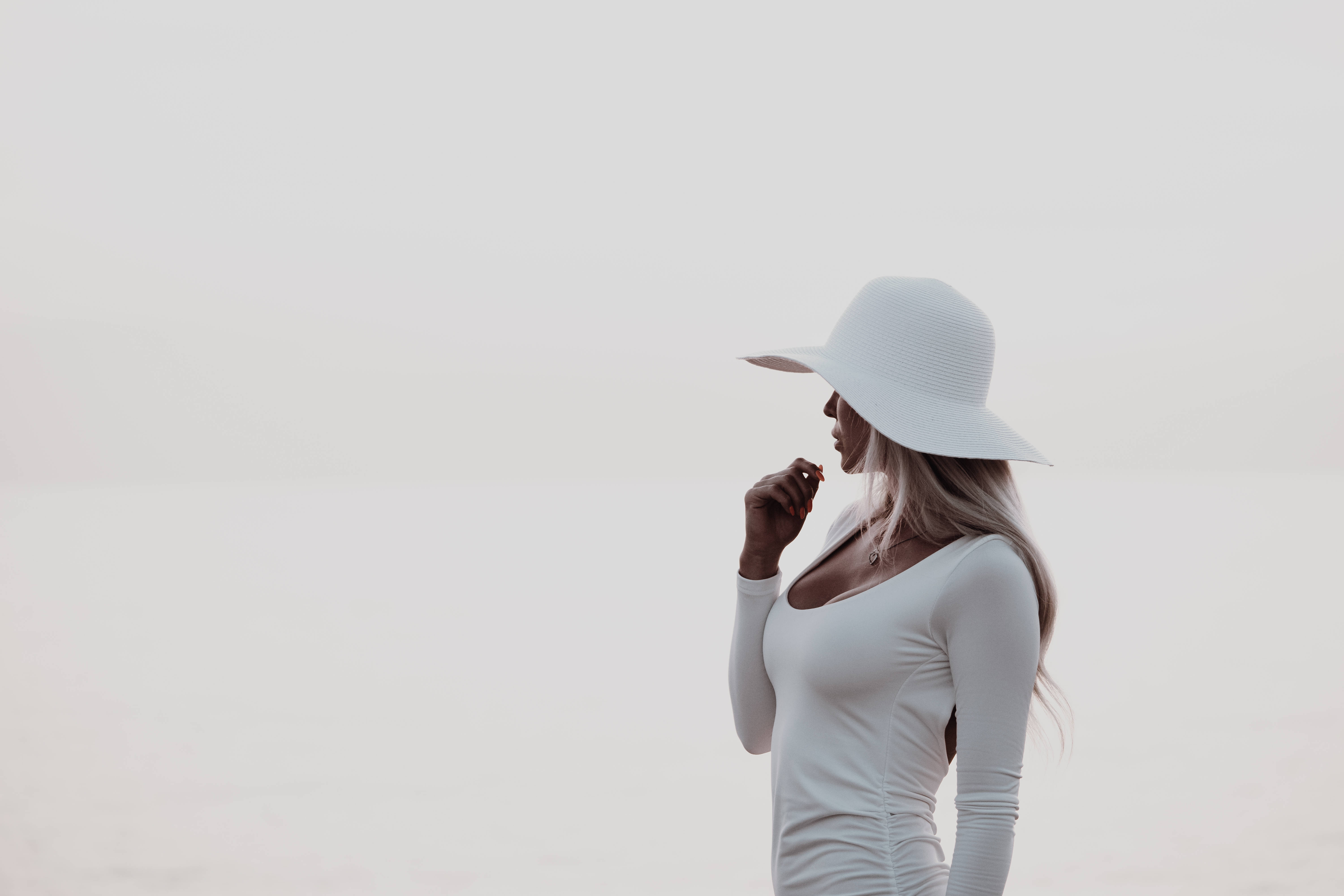 簡約白色美學 桌布 壁紙 — 一位戴白色帽子和白色上衣的女性側身站立，背景柔和朦朧，手靠近嘴邊。