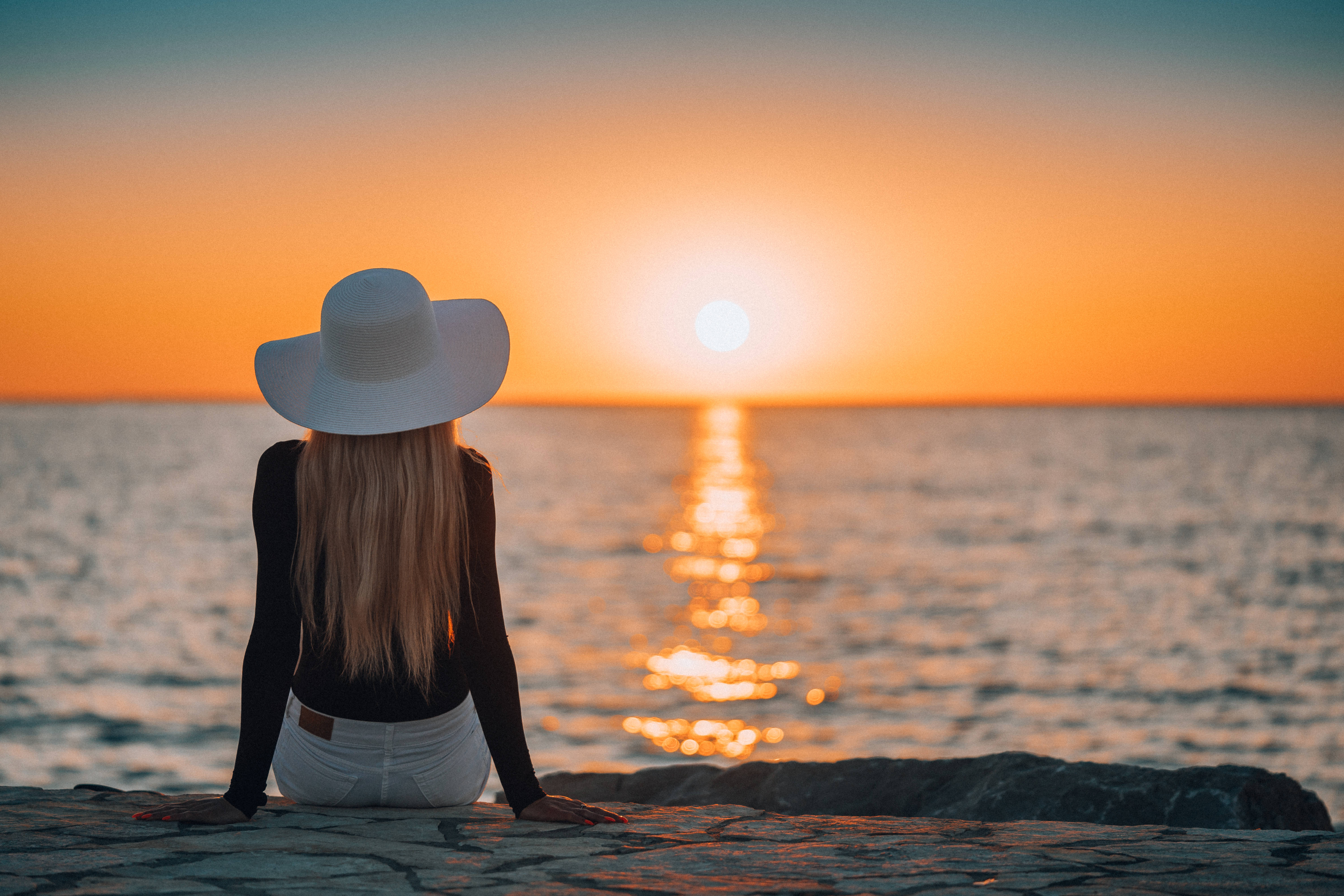 海洋 桌布 壁紙 — 一位戴白色帽子的女士坐在石台上，觀賞夕陽沉入海洋，金色光芒映照在水面上。