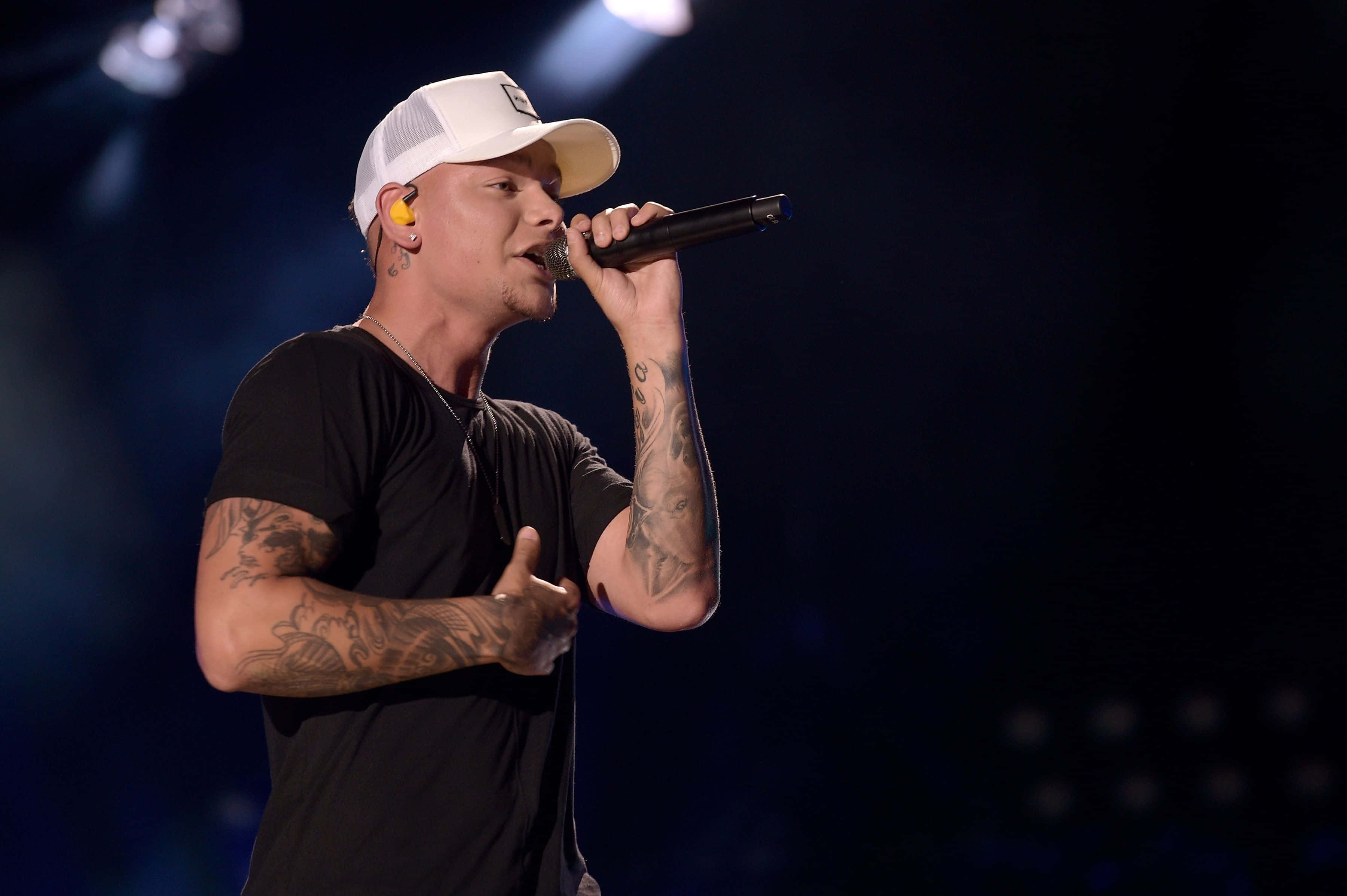 Kane Brown 桌布 壁紙 — 一位戴白色鴨舌帽、有刺青的男歌手在舞台燈光下熱情地對著麥克風演唱。