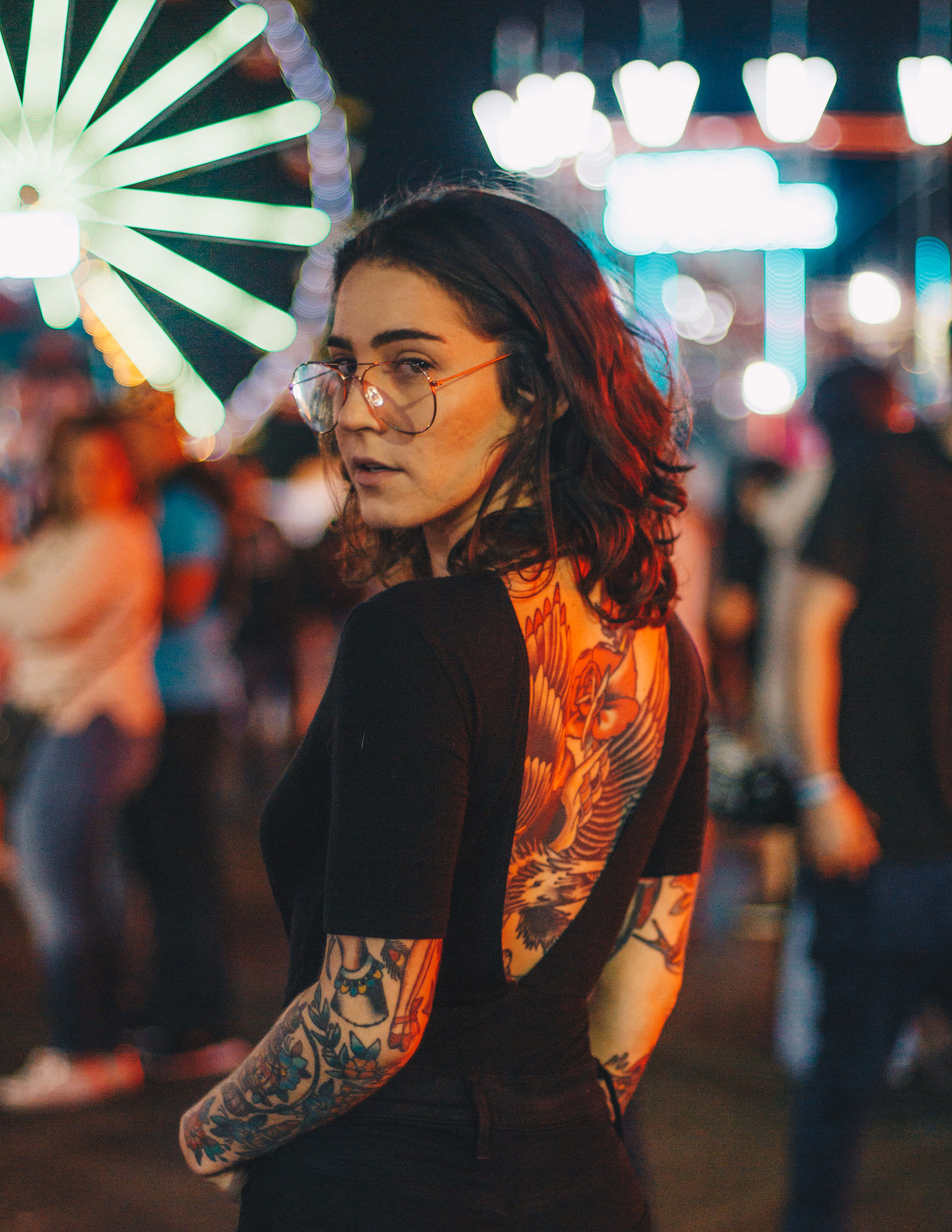 Tattoo Girl 桌布 壁紙 — 一位戴眼鏡、擁有複雜刺青的女性在夜晚嘉年華的閃爍燈光下轉向鏡頭。