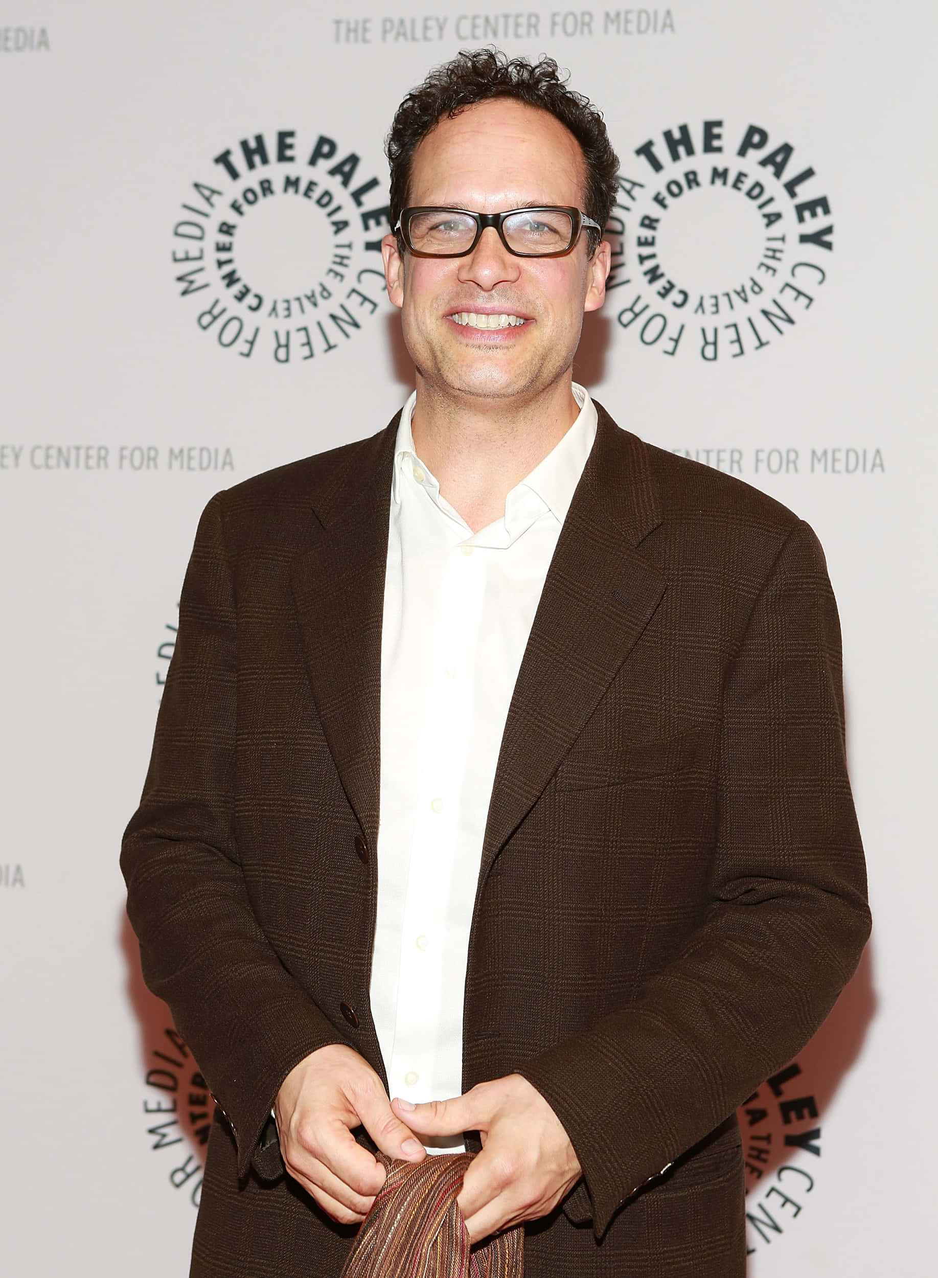 Diedrich Bader 桌布 壁紙 — 一位戴眼鏡、深色頭髮的微笑男士，在媒體中心活動中穿著棕色格紋西裝外套，內搭白襯衫。