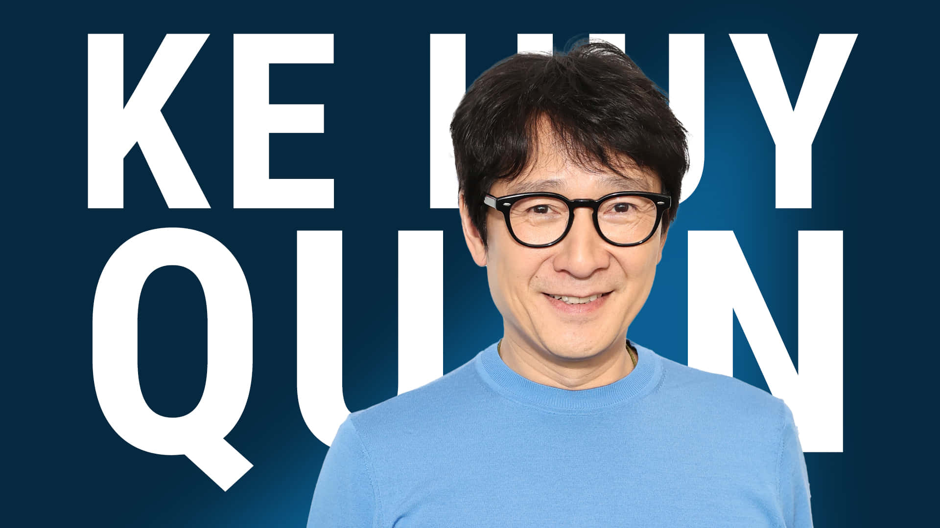 Ke Huy Quan 桌布 壁紙 — 一位戴眼鏡、深色頭髮的微笑男士，身穿藍色襯衫，背景為深藍色，並有大型白色文字拼出「KE HUY QUAN」。