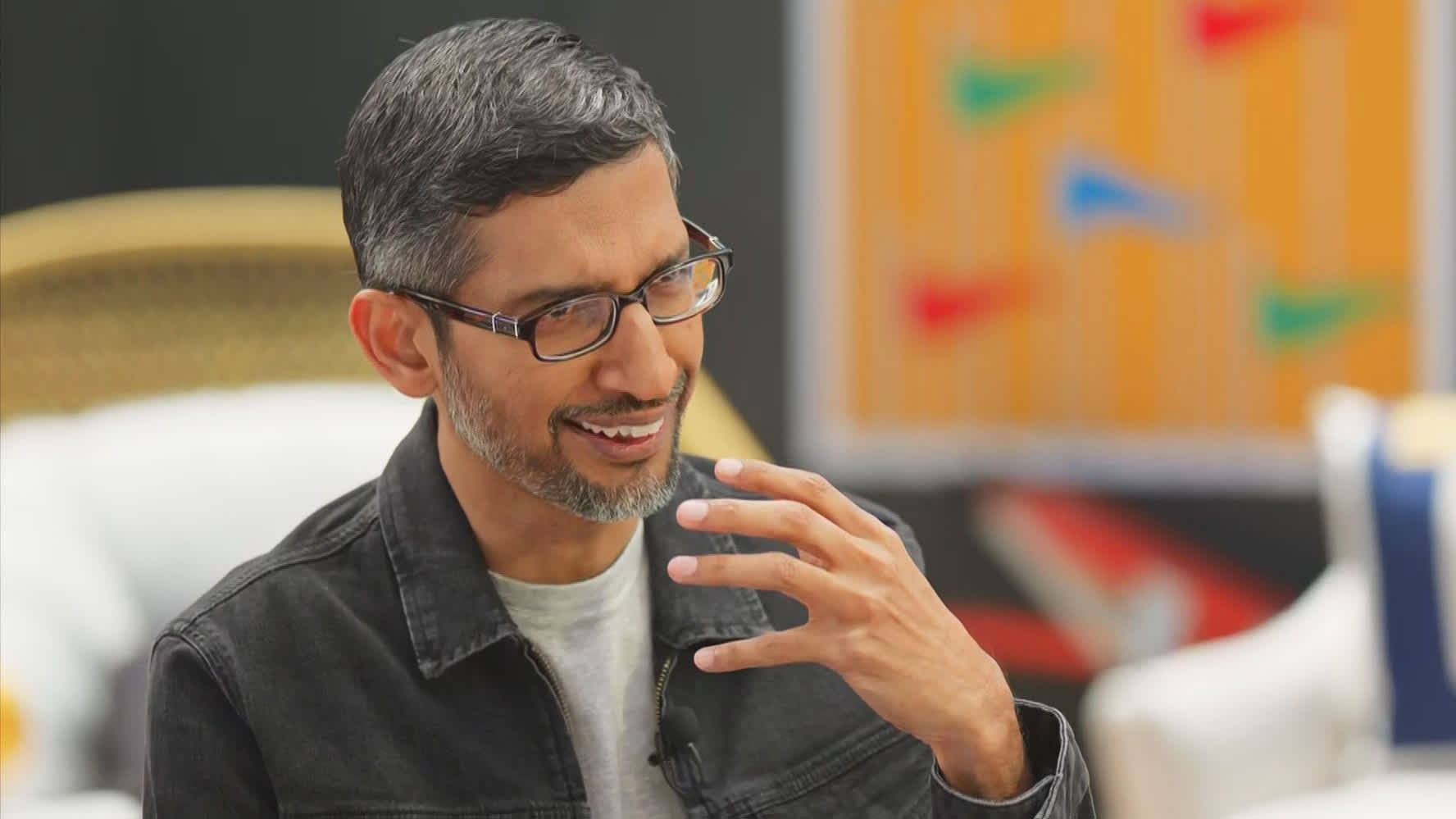 google 桌布 壁紙 — 一位戴眼鏡、灰白頭髮的男子在現代室內環境中微笑並做手勢。