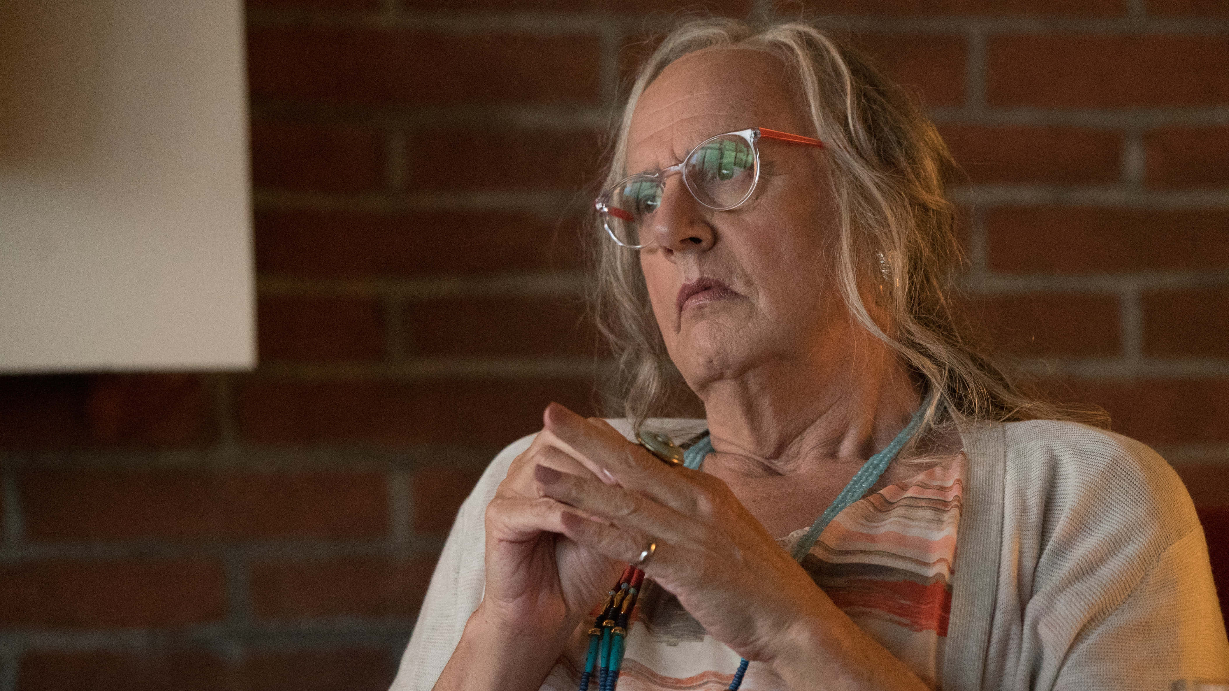 jeffrey tambor 桌布 壁紙 — 一位戴眼鏡、灰白頭髮的老年女士靠著紅磚牆沉思地坐著，雙手交握在胸前。