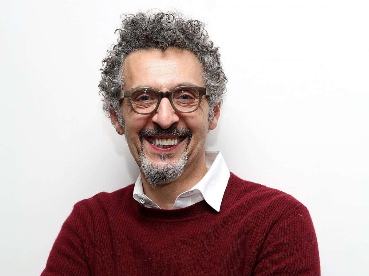 John Turturro 桌布 壁紙 — 一位戴眼鏡、灰色捲髮的開朗男士，穿著紅色毛衣，在純白背景前溫暖微笑。