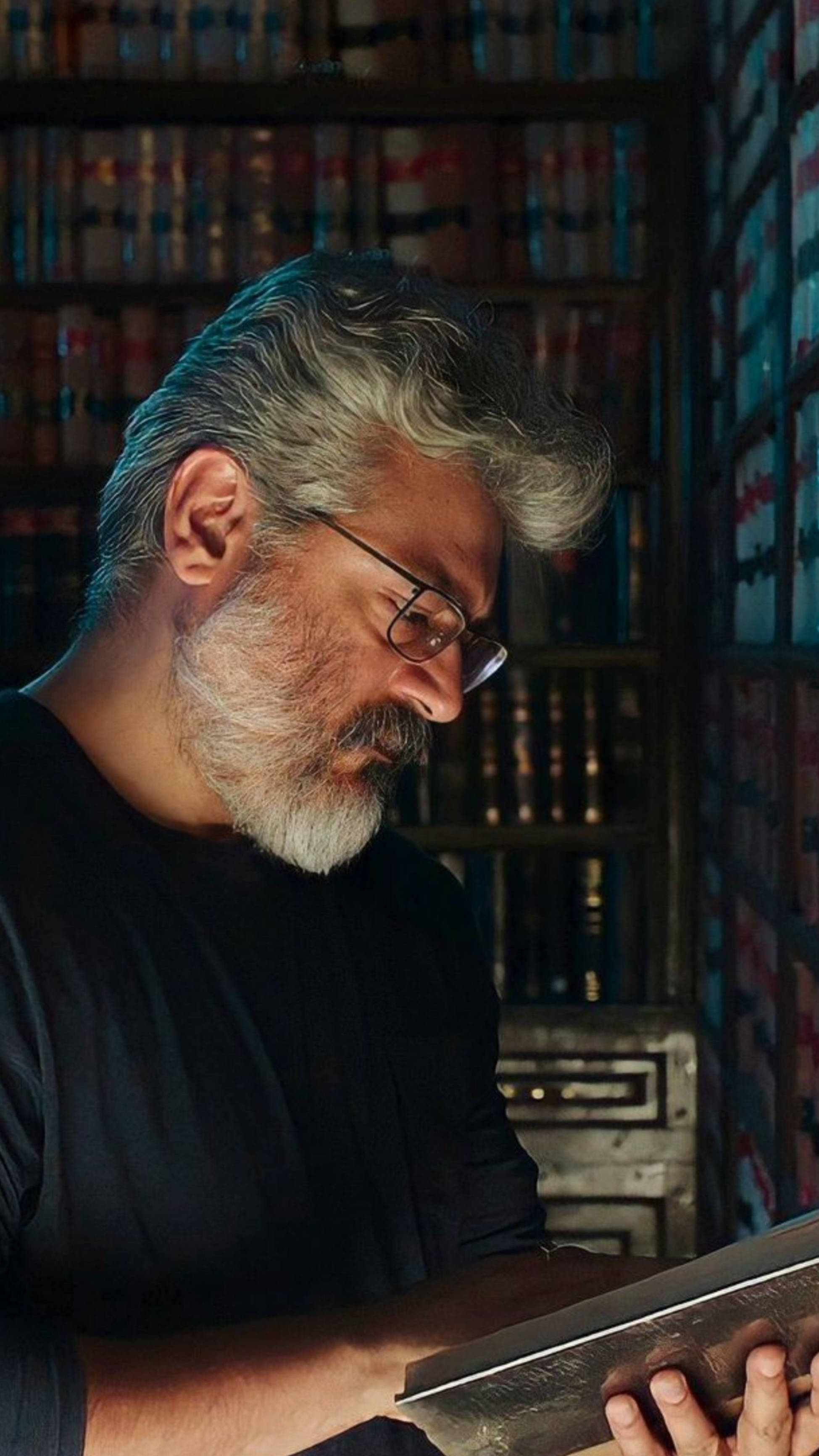 actor 桌布 壁紙 — 一位戴眼鏡、灰髮灰鬍鬚的男子在昏暗的圖書館中，被高聳的書架環繞，專注地閱讀一本書。