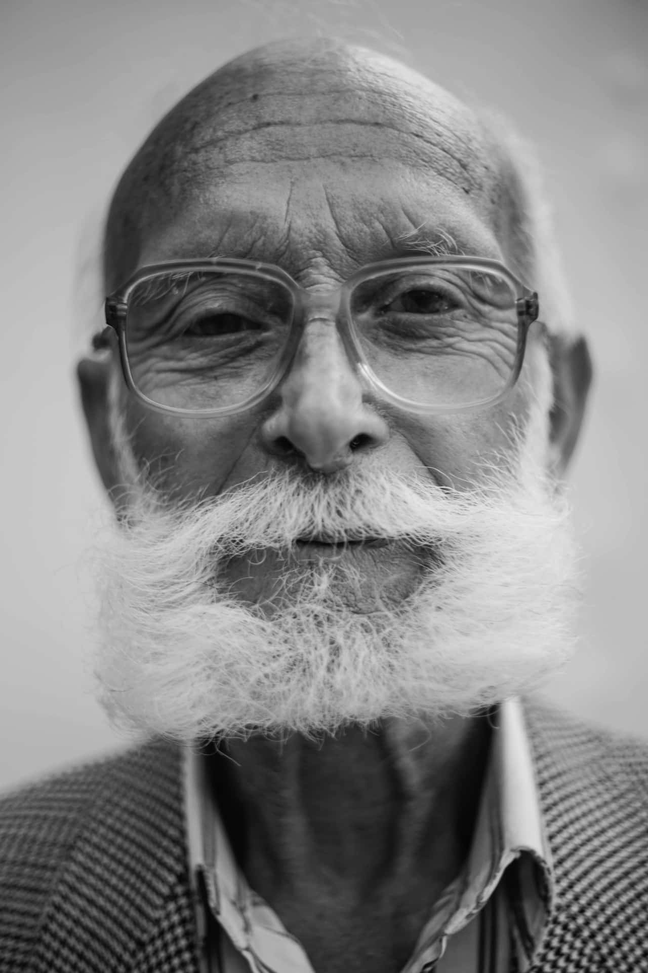 黑白 桌布 壁紙 — 一位戴眼鏡、留著白色鬍子的長者，對著鏡頭溫柔微笑的特寫黑白肖像。
